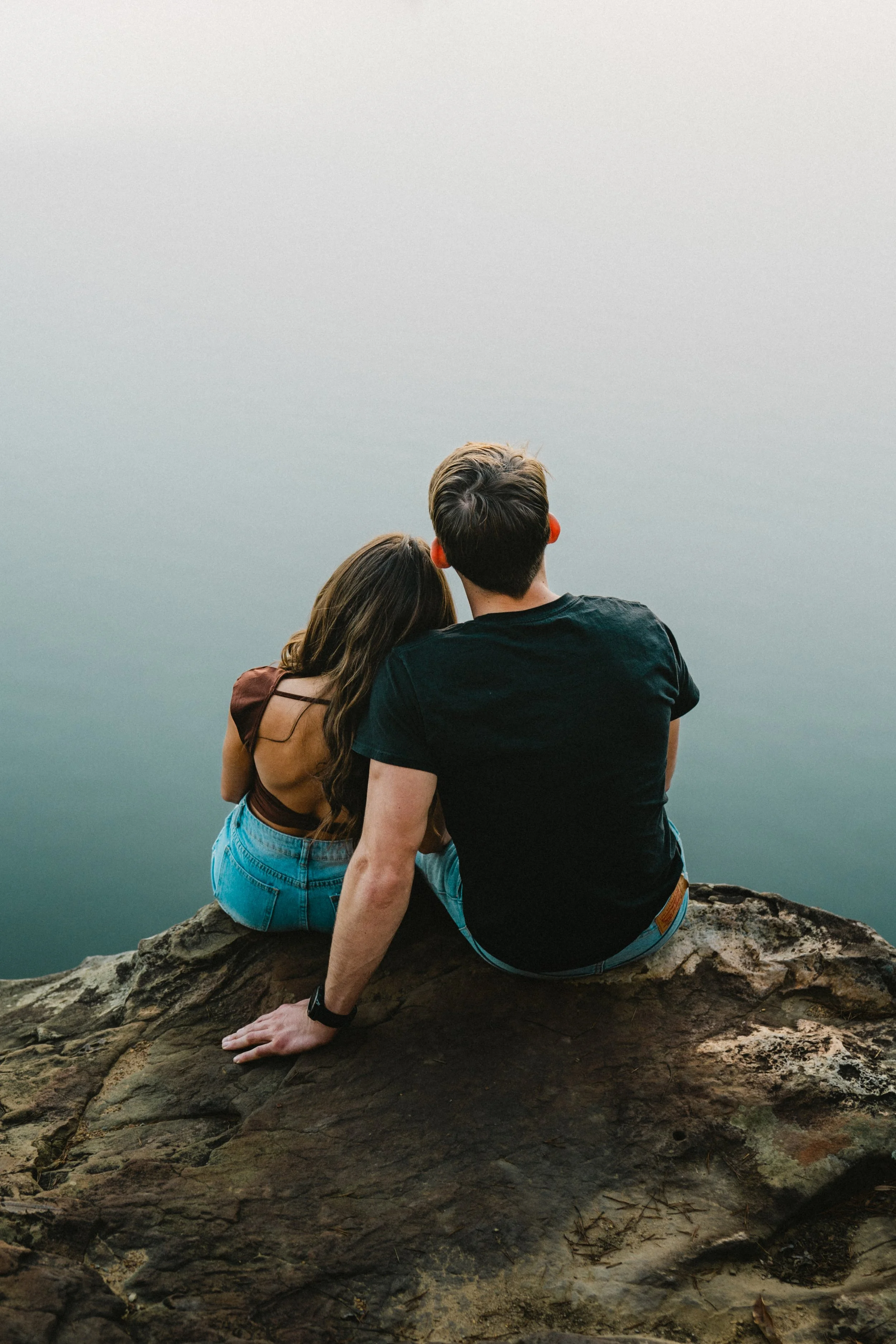 A man and woman sitting on a rocky ledge overlooking a misty body of water, with their backs facing the camera.