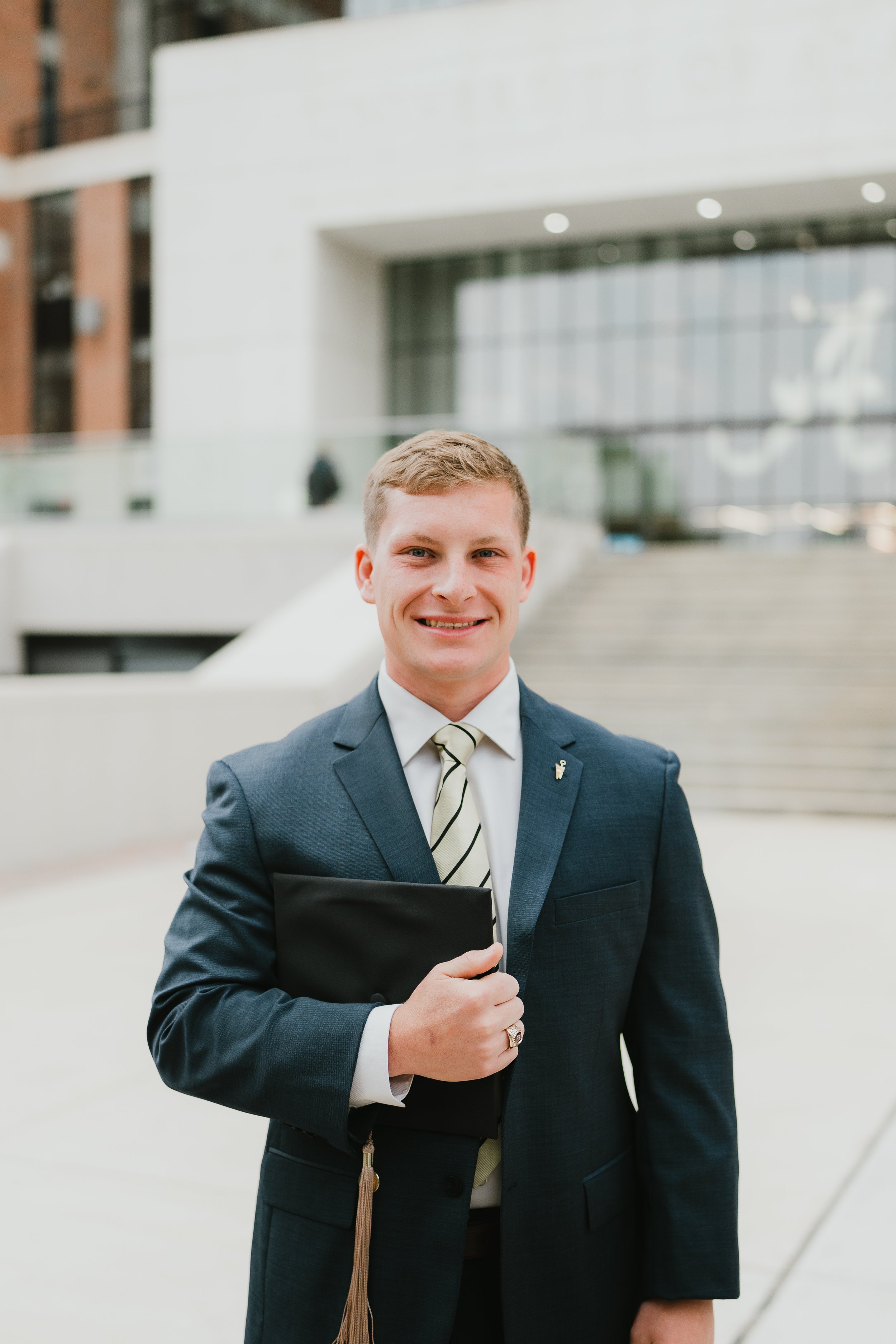 A young man in a suit holding a folder and smiling standing outside a modern building.