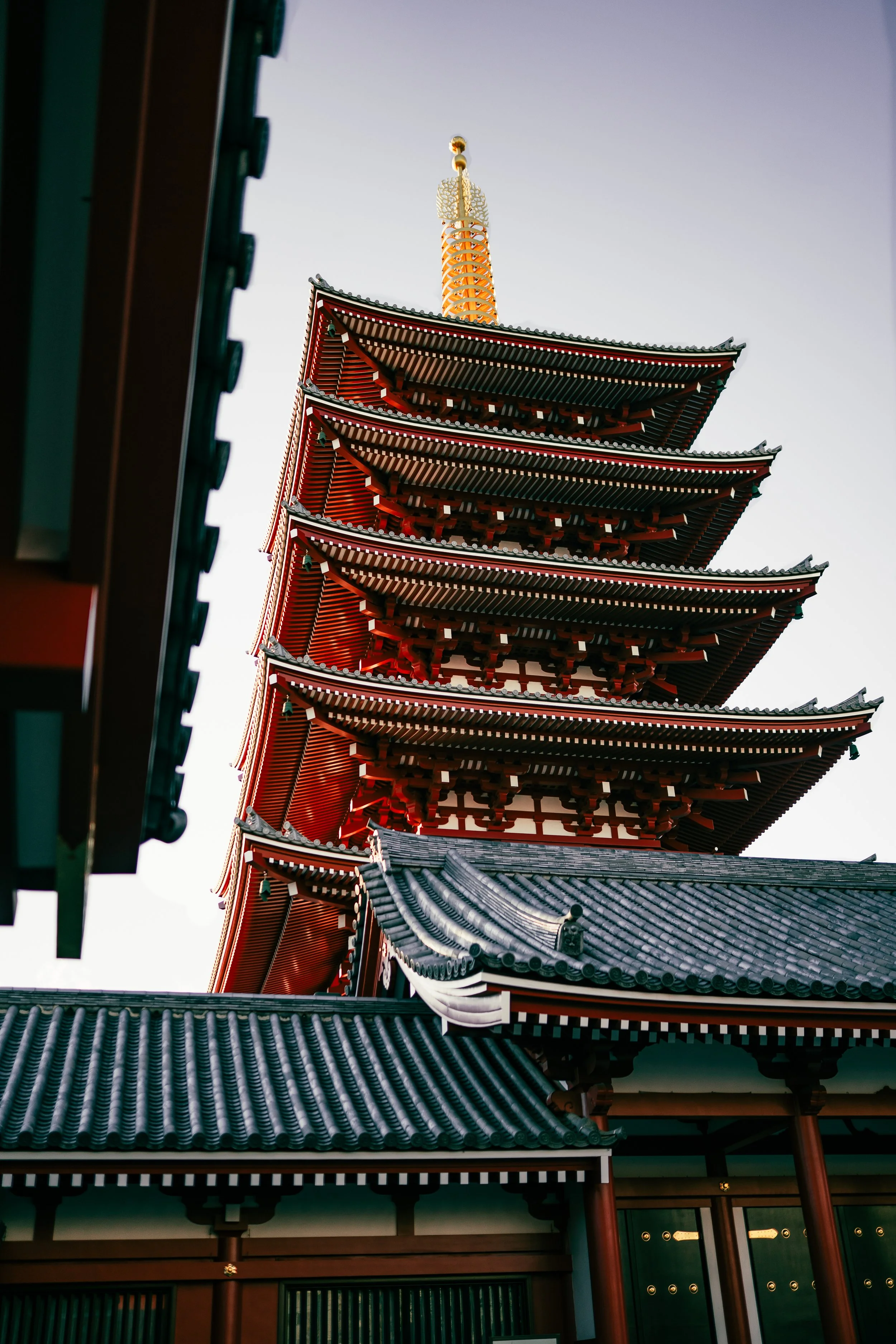 A traditional Japanese pagoda with a multi-tiered roof, ornate eaves, and a tall spire topped with gold decorations, set against a clear sky.