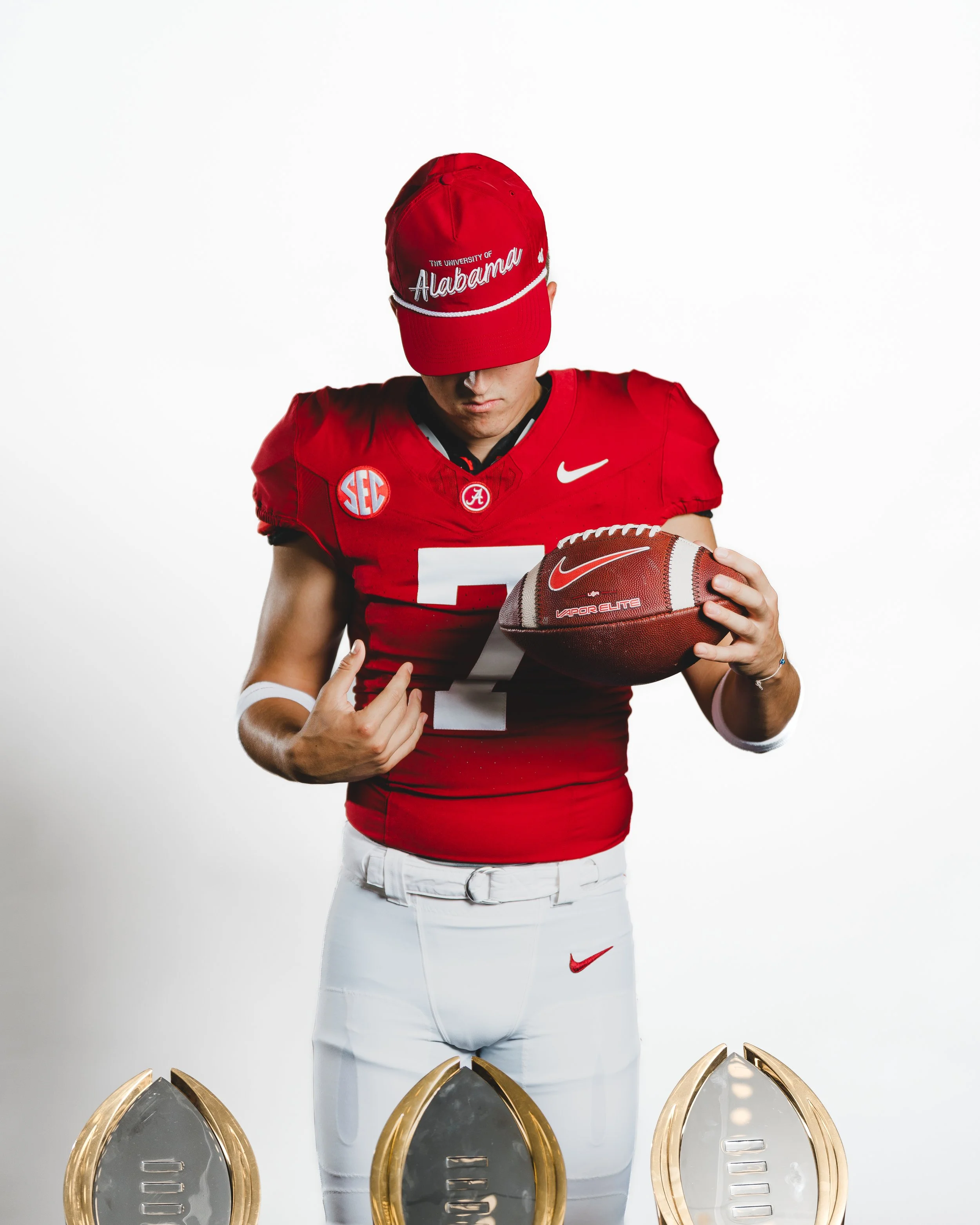A football player in a red jersey and white pants holds a football and looks down, wearing a red Alabama cap. The background is plain white with three gold and silver trophies or awards at the bottom of the image.