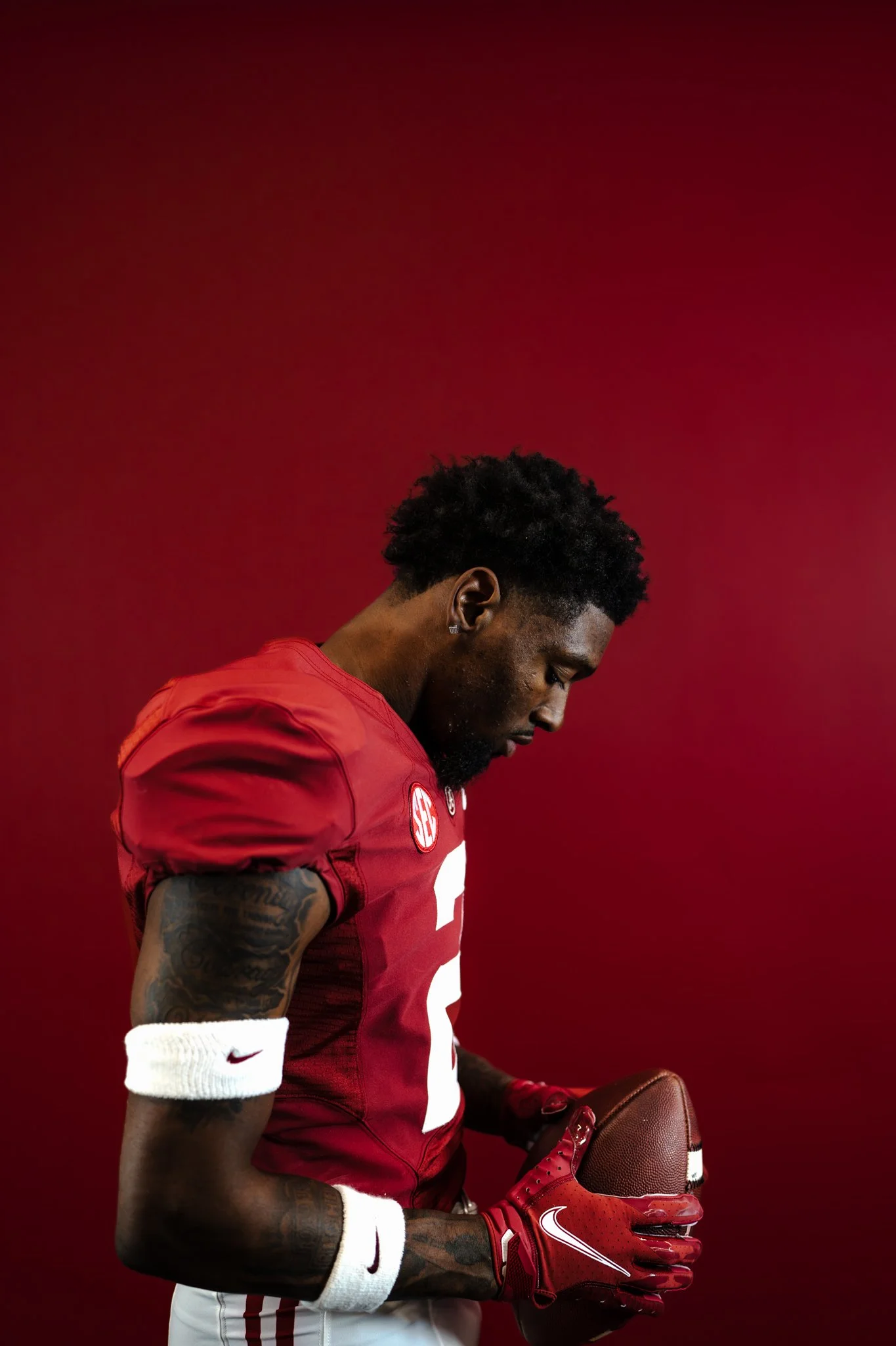 A young athletic man with tattoos on his arm wearing a red sports jersey, red gloves, and white wristbands, holding a football against a solid red background. He is looking downward.