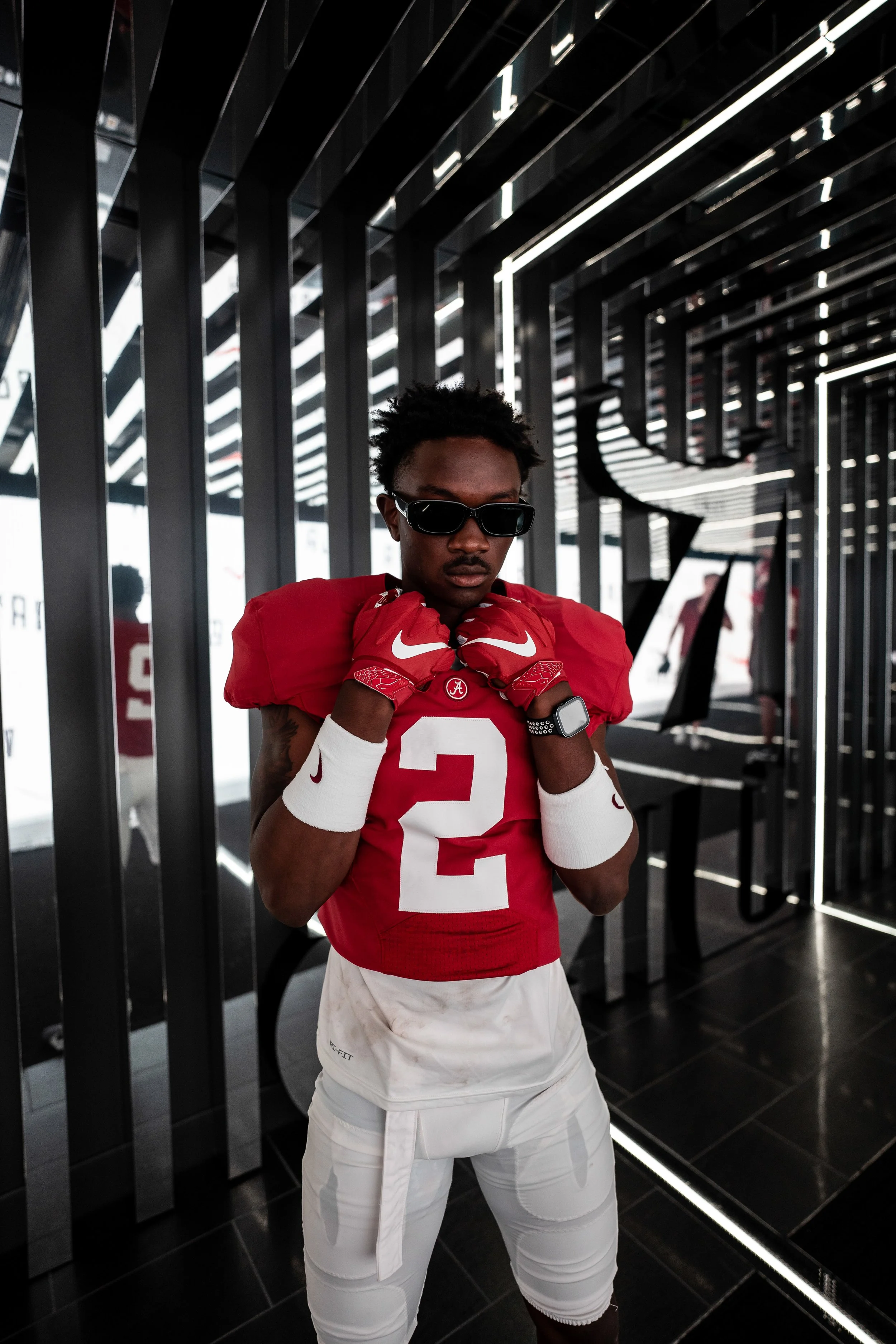 A football player in a red jersey with the number 2, wearing sunglasses, white pants, and red gloves, standing in a modern, black, metallic, tunnel-like structure with geometric lighting overhead.