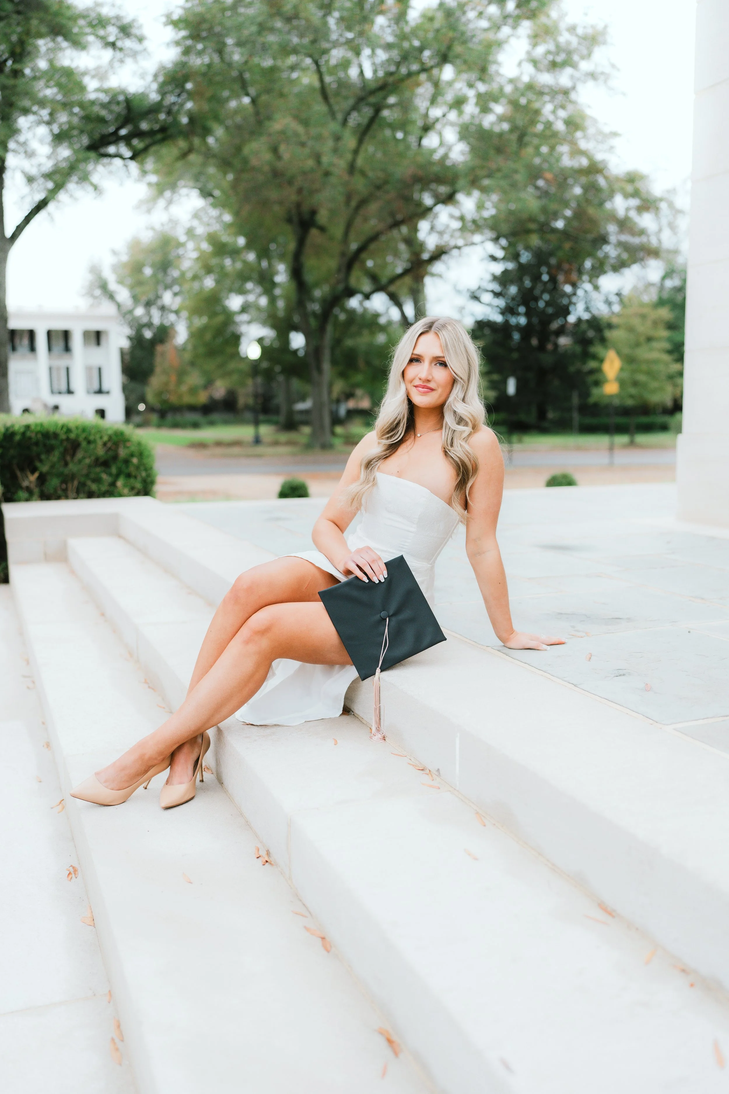 A young woman in a white dress sitting on white steps outdoors, holding a black graduation cap, with trees and a building in the background.
