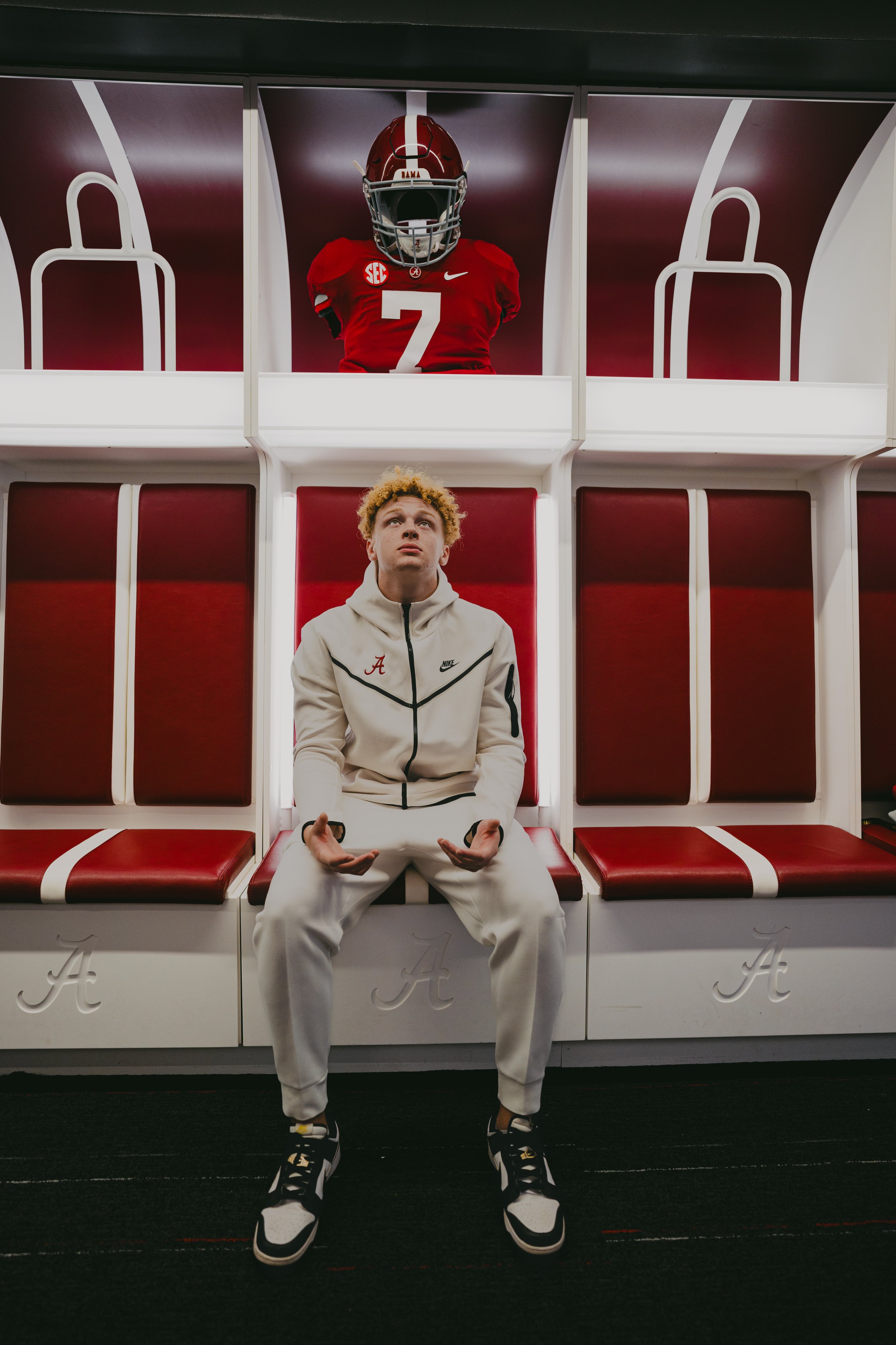 A young man with curly blonde hair wearing a white hoodie and white pants sitting on a bench in a sports locker room. Behind him, an Alabama football helmet and jersey with the number 7 are displayed in a glass case.