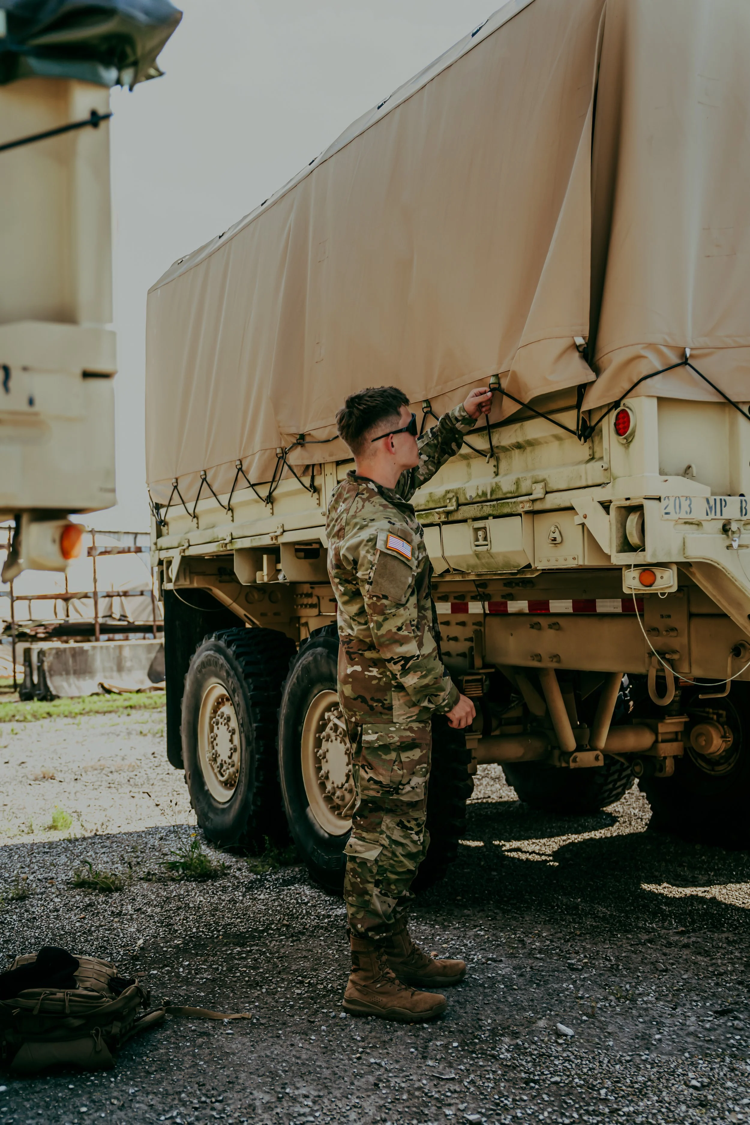 A soldier in camouflage uniform standing next to a military vehicle with a covered cargo area, adjusting a strap or handle. The ground is gravel with some grass, and backpacks are on the ground nearby.