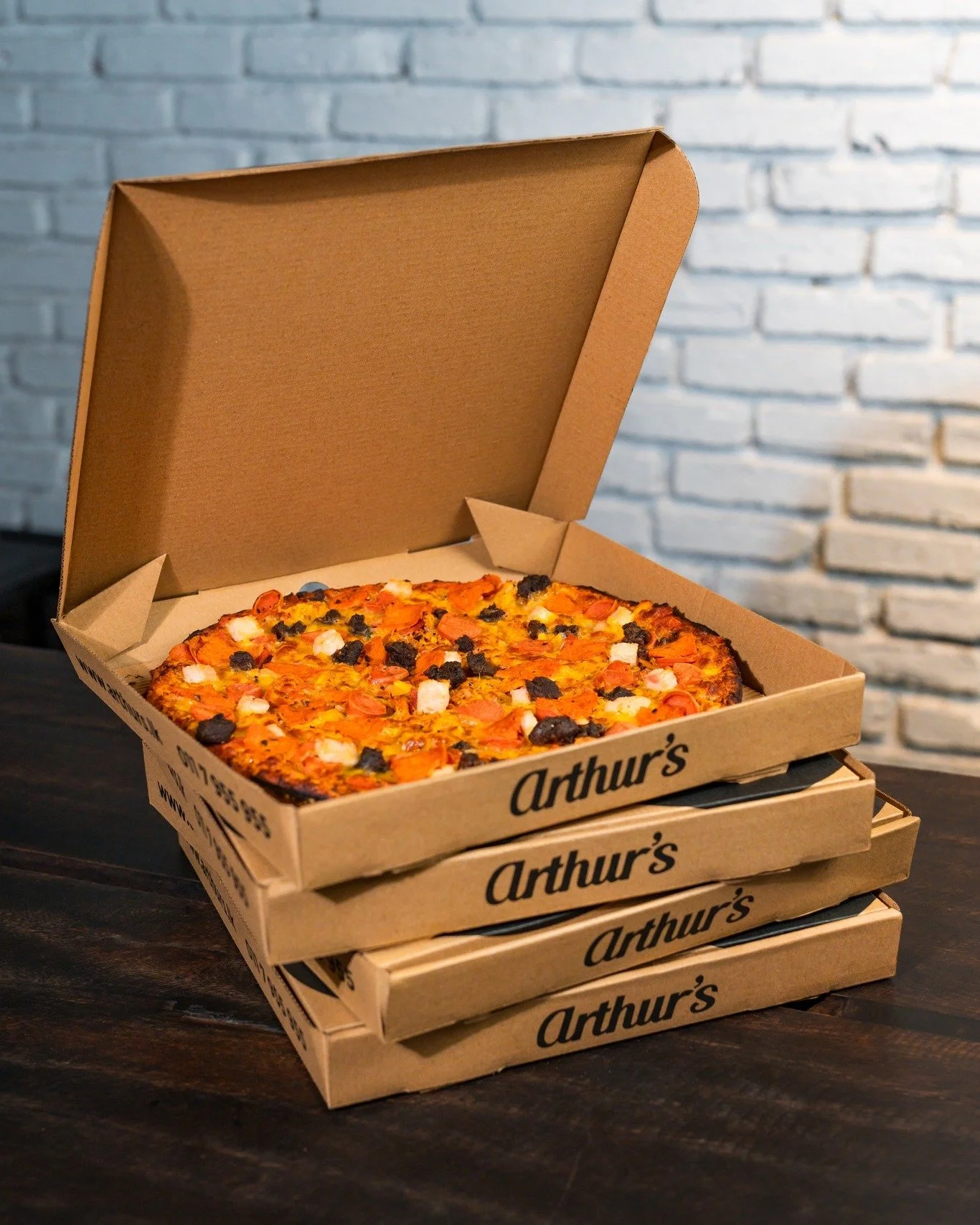 Stack of four pizza boxes labeled "Arthur's" with an open pizza box on top showing a pizza with various toppings, placed on a dark wooden surface against a brick wall background.