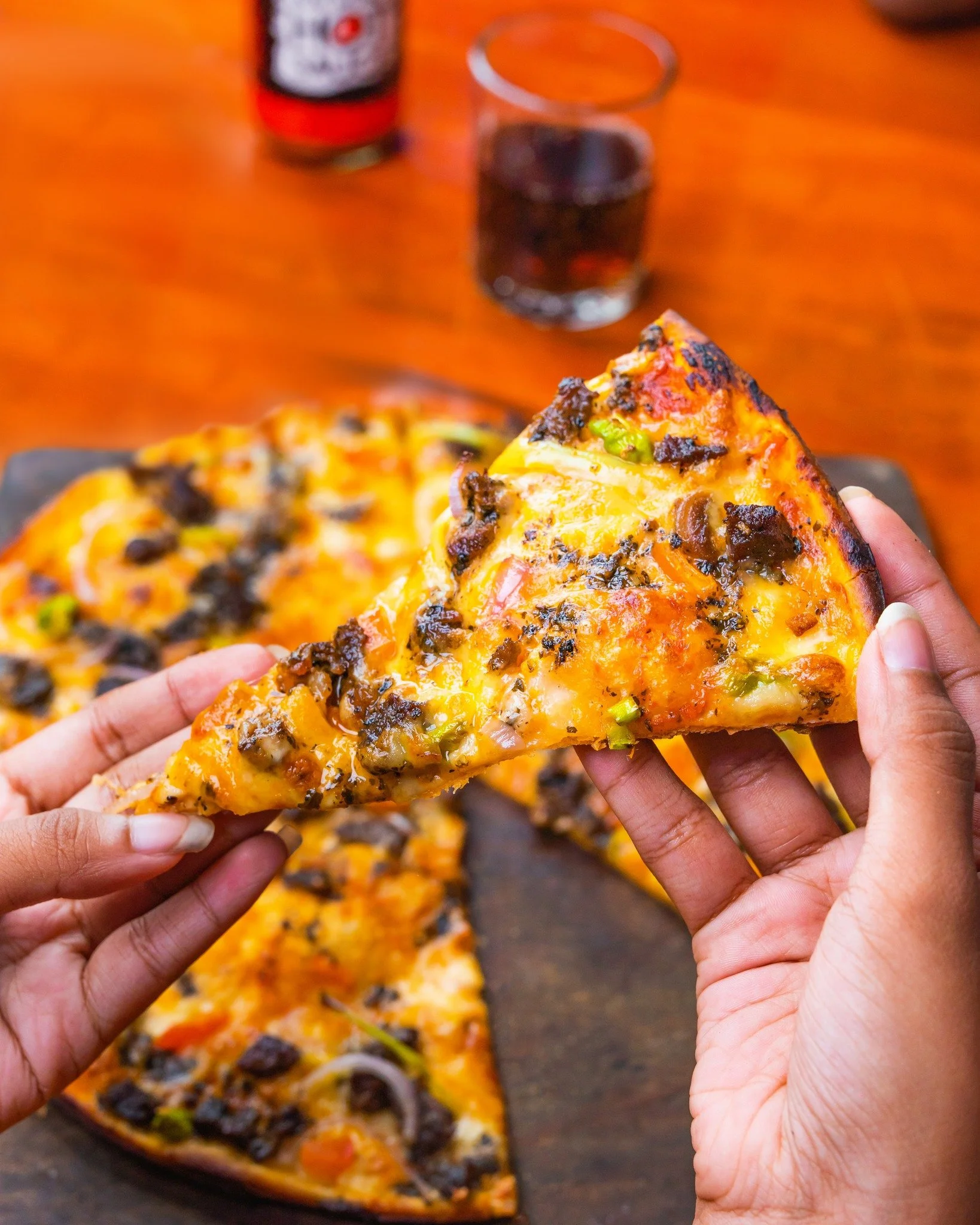 Two hands holding a slice of pizza with cheese, mushrooms, green peppers, and black olives, with a pizza in the background and a glass of dark soda and a soda can on a wooden table.