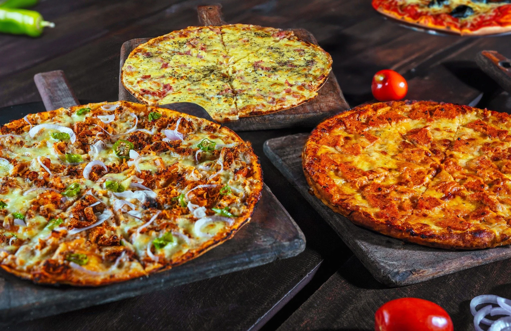 Several pizzas on black slabs, featuring cheese, meats, vegetables like onions and peppers, and various toppings, on a dark wooden surface with small tomatoes and chili peppers nearby.