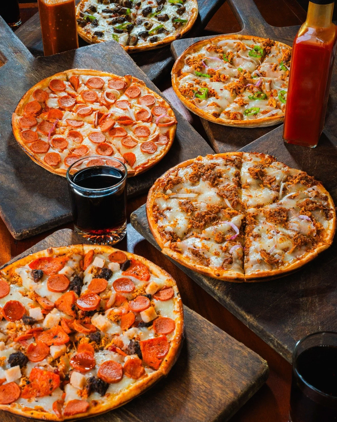 Several pizzas with various toppings such as pepperoni, sausage, and ground meat, along with bottles of ketchup and soda on a dark wooden table.