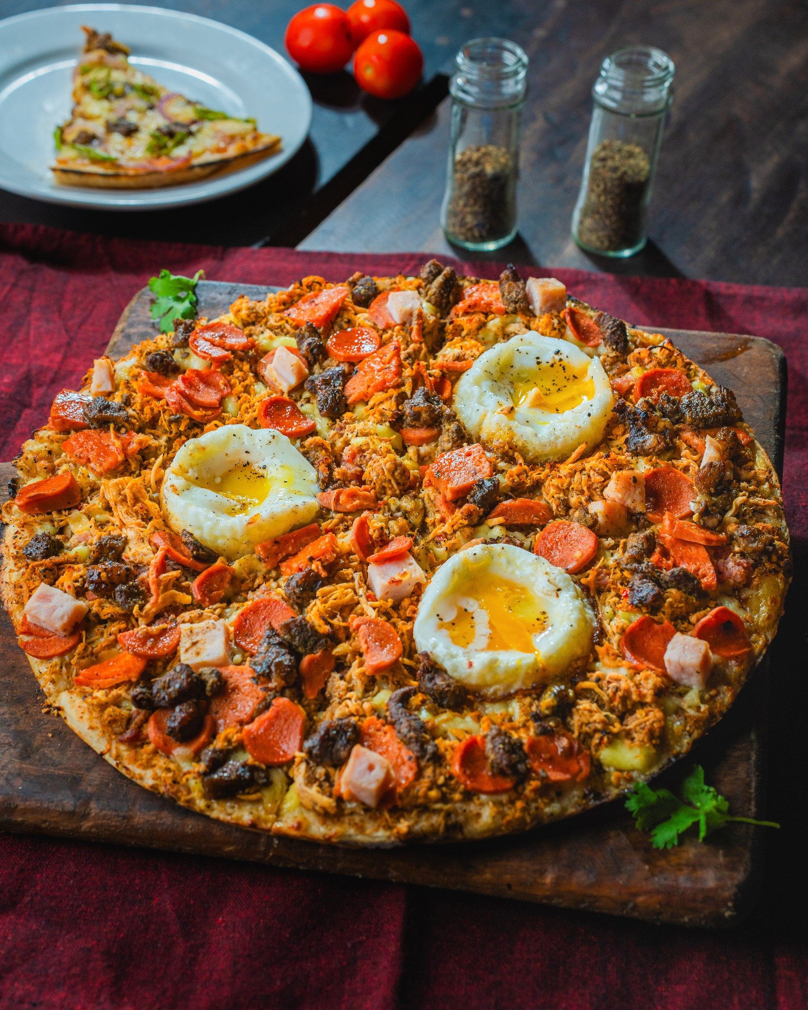 Close-up of a pizza with sunny side up eggs, pepperoni, sausage, and vegetables on a wooden serving board, with tomatoes and spice jars in the background.