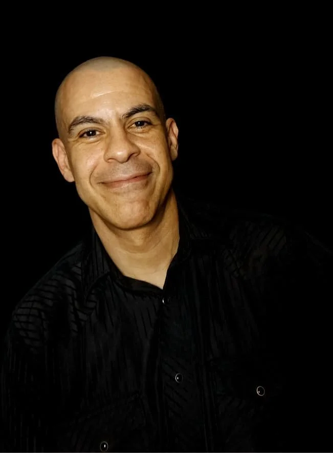 Hombre sonriendo, piel clara, cabeza calva, con camisa negra, fondo negro.