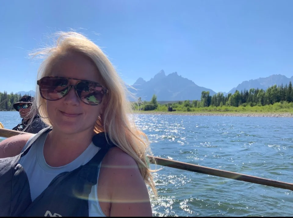 Woman with long blonde hair, wearing sunglasses and a light-colored top, sitting on a boat with mountains and a forested shoreline in the background, on a sunny day.