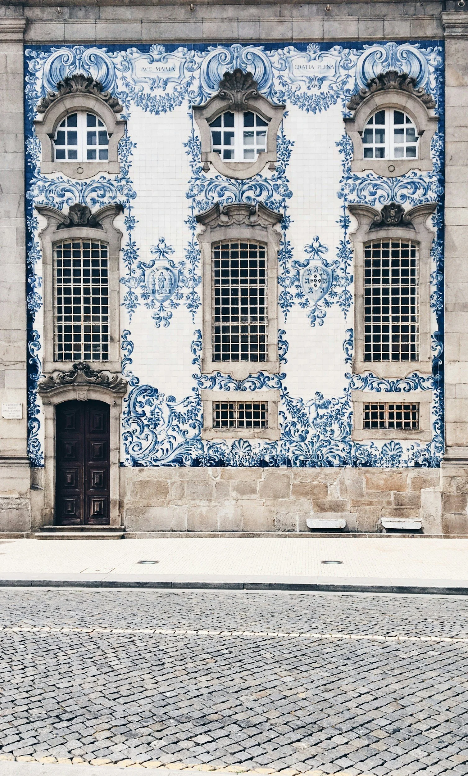 Facade of a building decorated with traditional blue and white ceramic azulejos tiles, featuring windows with decorative stone frames, a central plaque, and a wooden door, situated on a cobblestone street.