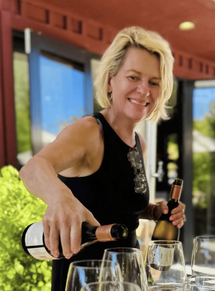 A woman with short blonde hair pouring wine into glasses at a table outdoors.