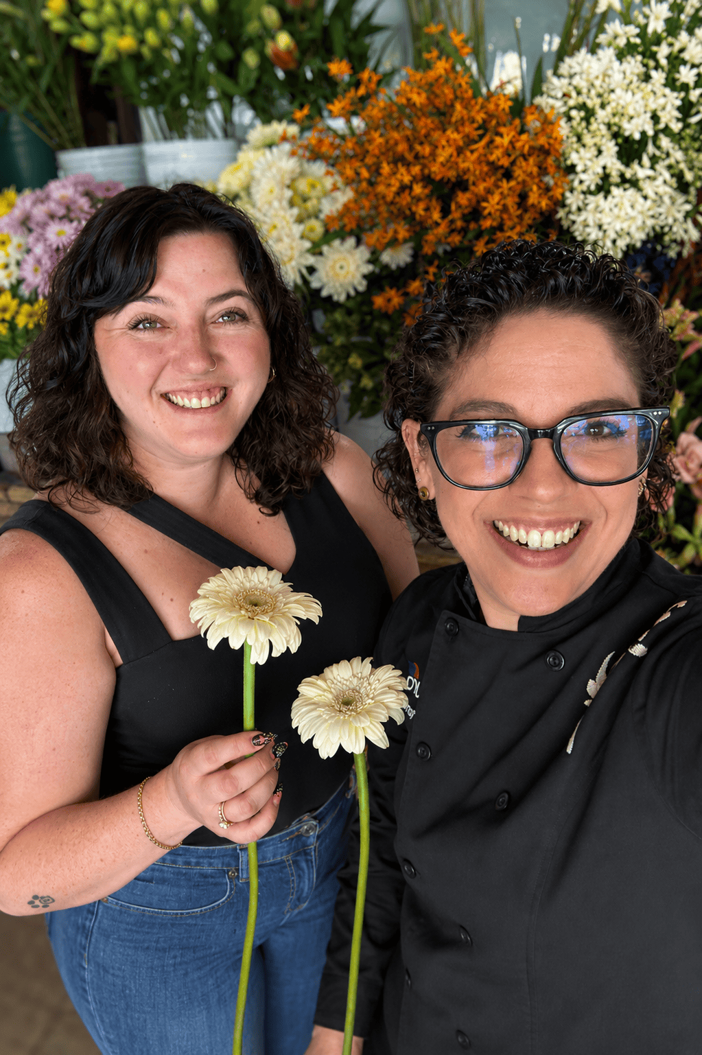 Two women smiling, holding white gerbera daisies, surrounded by colorful flowers in the background.