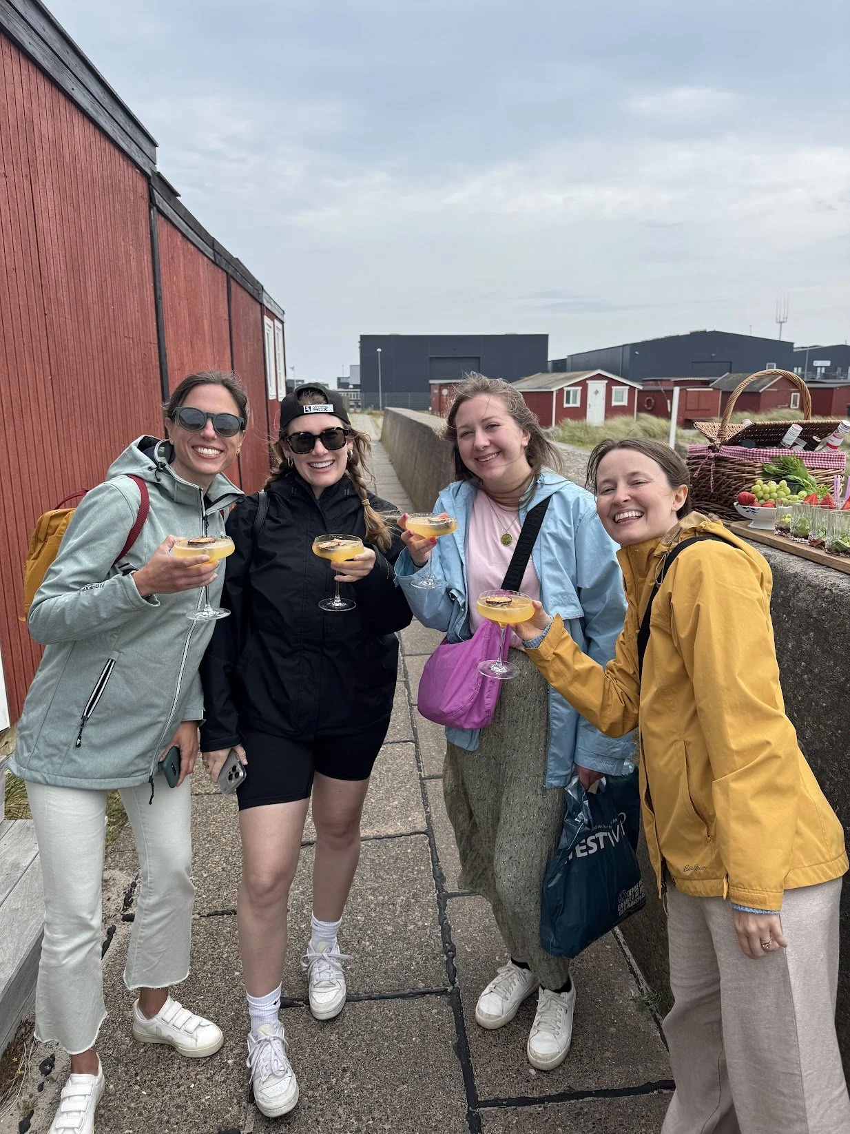 Four women wearing jackets and sunglasses, smiling and holding cocktails, standing on a street near a red building and a table with fruits and a basket of drinks.