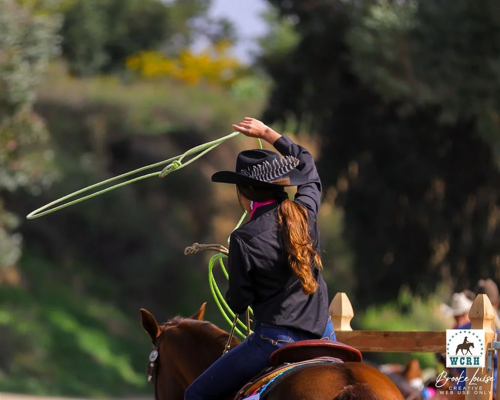 A woman riding a horse during a rodeo event, wearing a black hat and jacket, twirling a lasso in the air.