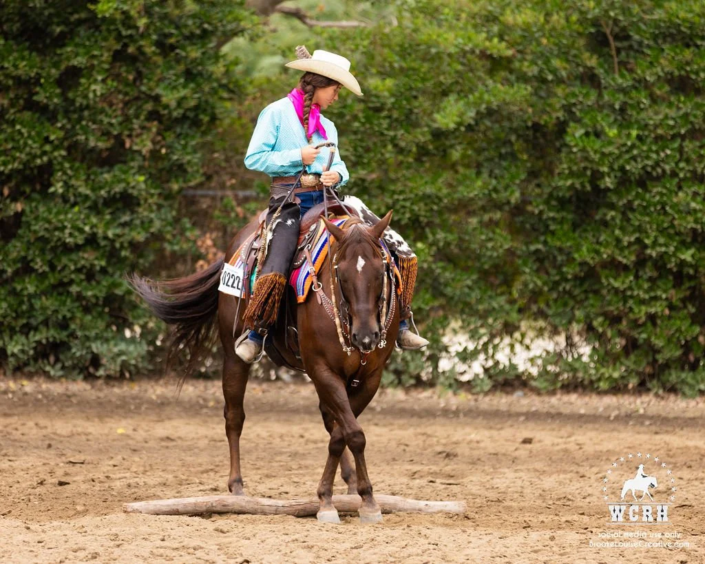 A young woman riding a horse on a dirt arena, surrounded by green bushes, wearing a cowboy hat, a turquoise button-up shirt, pink neckerchief, and western gear.