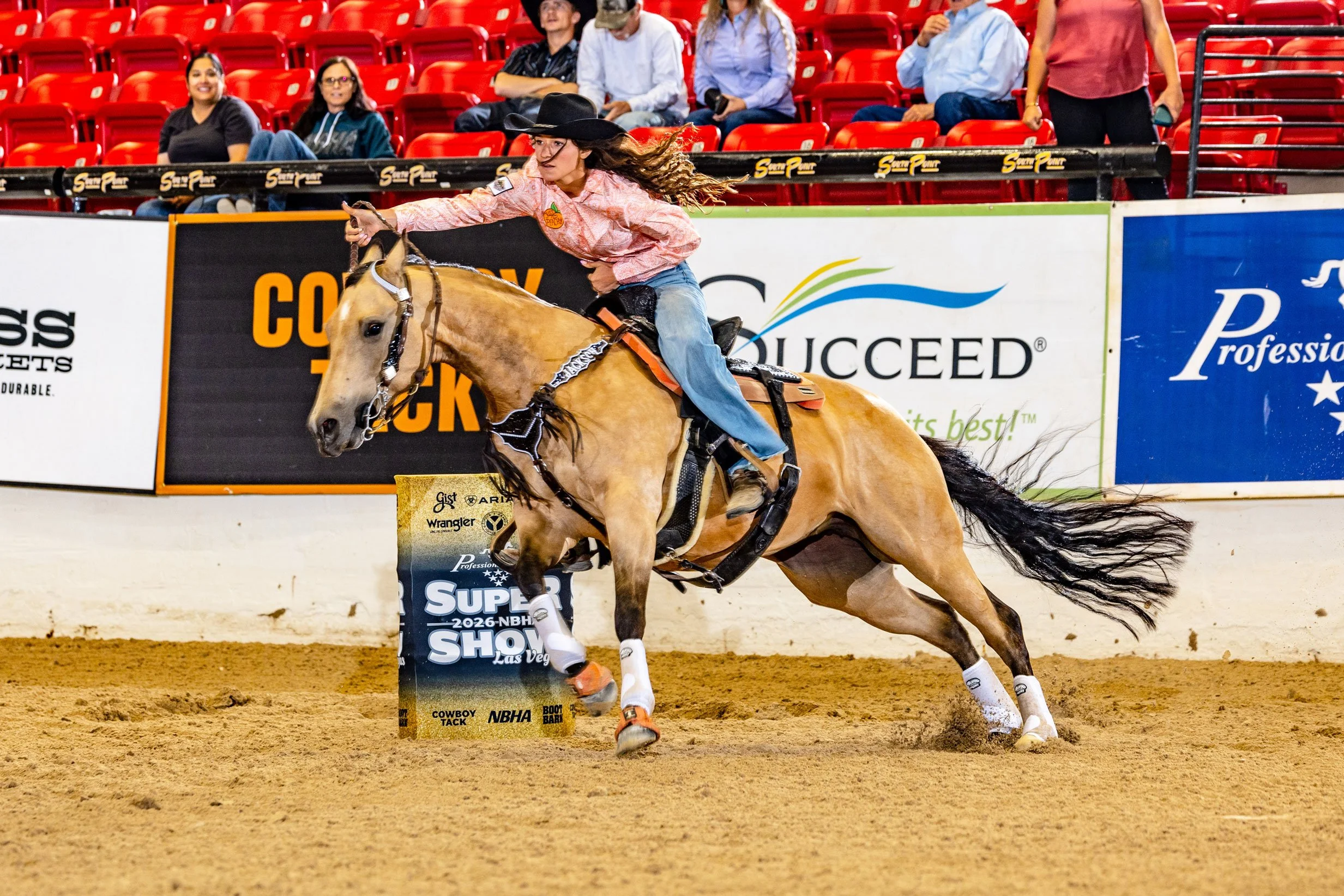 A woman dressed in Western attire riding a bucking horse in an indoor rodeo arena, with spectators seated in red seats in the background.