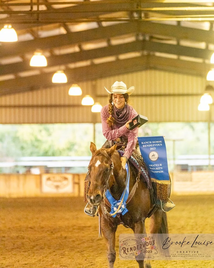 Young woman wearing a cowboy hat and plaid shirt riding a horse inside an indoor riding arena, holding a black box and smiling, with a blue ribbon and a banner on the horse indicating an amateur championship win in 2023.