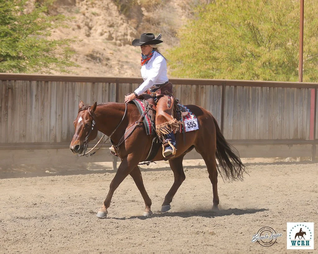 A woman riding a horse in a rodeo arena, wearing a cowboy hat, white shirt, and bandana, with a saddle and riding gear. There is a wooden fence and trees in the background.