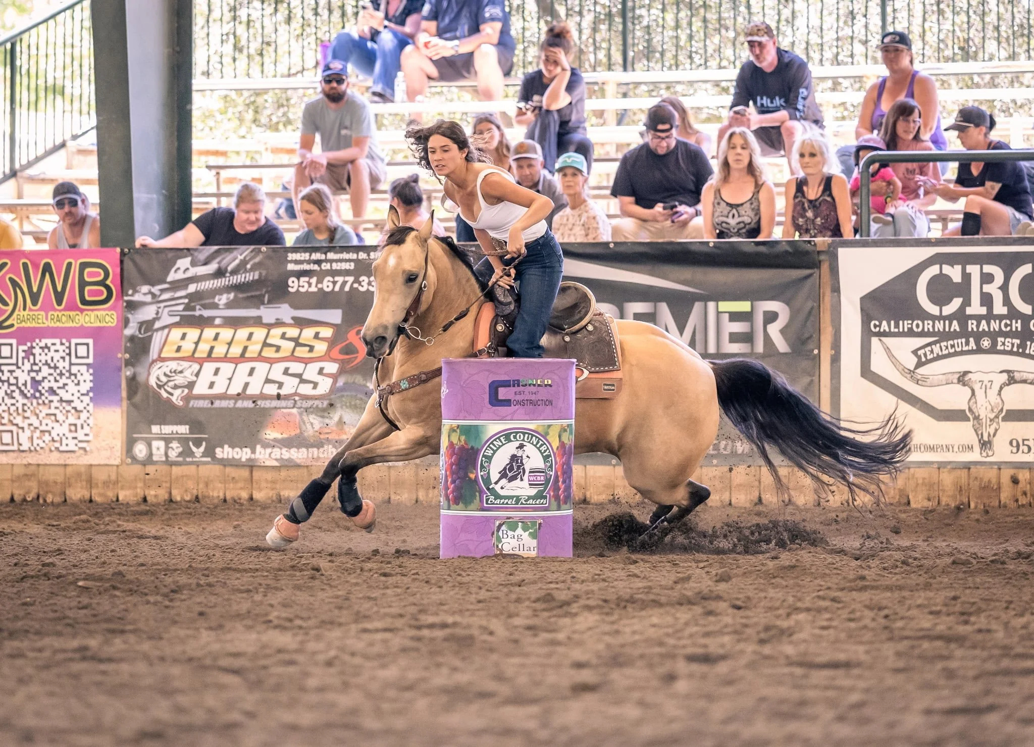 A woman riding a bucking horse during a barrel racing event at an indoor arena, with spectators watching from the stands.