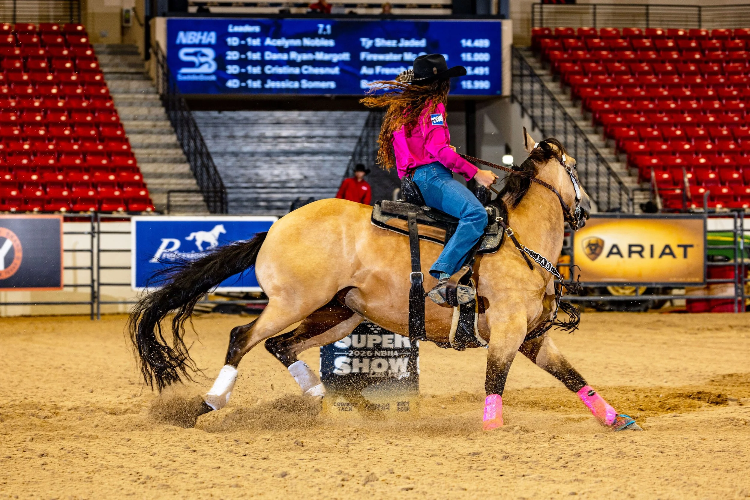 A female rodeo competitor riding a bucking horse in an indoor arena, wearing a pink shirt, jeans, and a black cowboy hat with long, flowing hair. The horse has a light-colored coat, black mane, and tail, and is wearing pink protective leg gear. In th