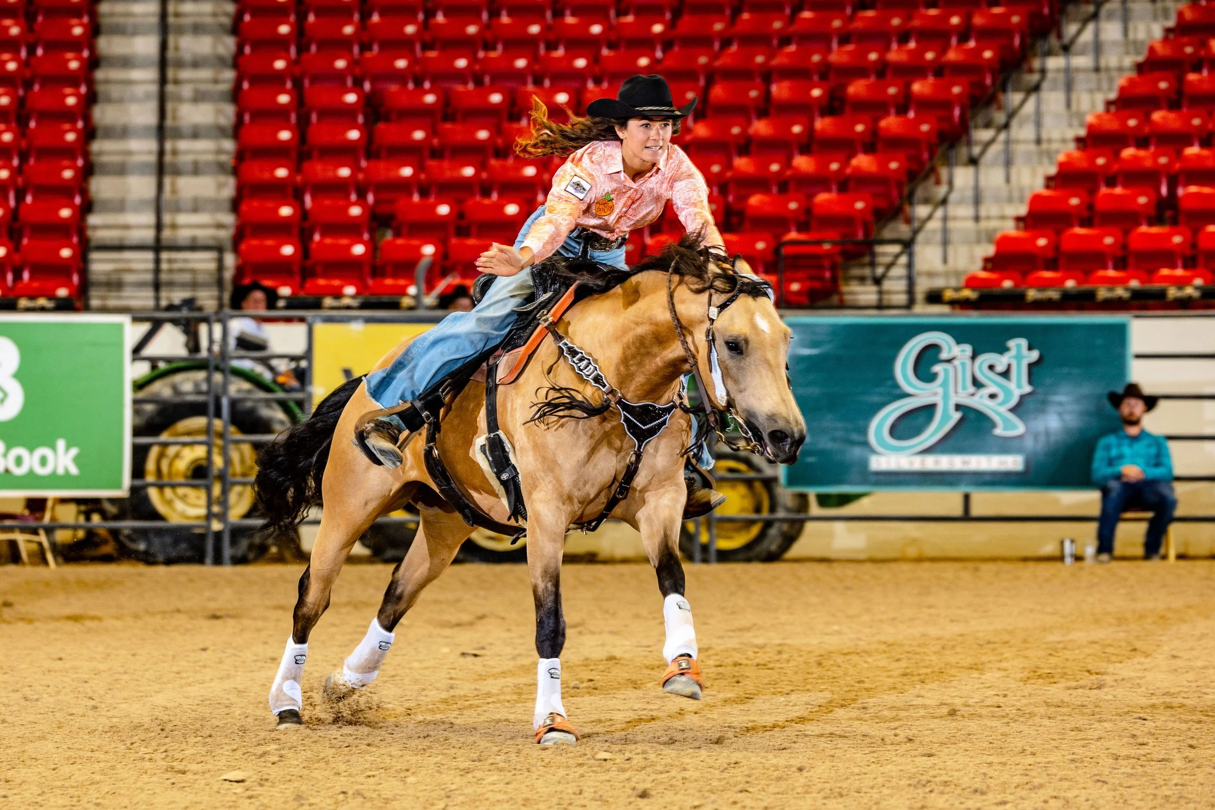 A woman riding a horse in an indoor arena, wearing a black cowboy hat, plaid shirt, and jeans, surrounded by red stadium seats.