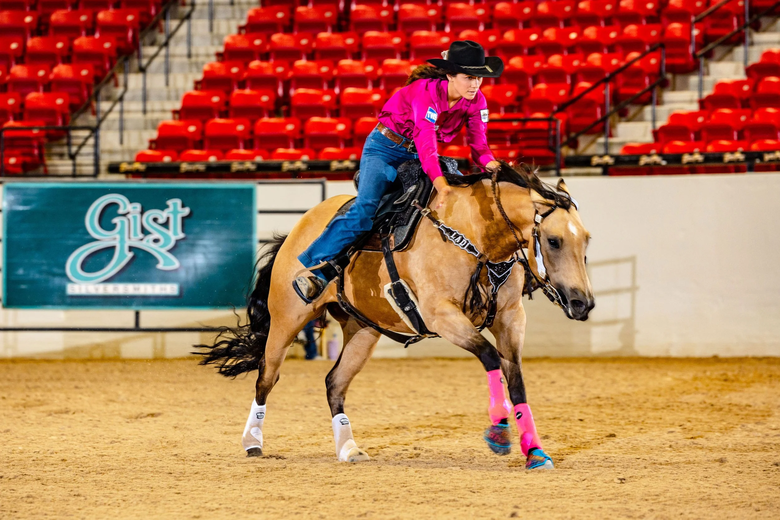 A female rodeo rider wearing a black hat, pink long sleeves, blue jeans, and pink protective gear on her legs, riding a bucking bronco inside an arena with red seats in the background.