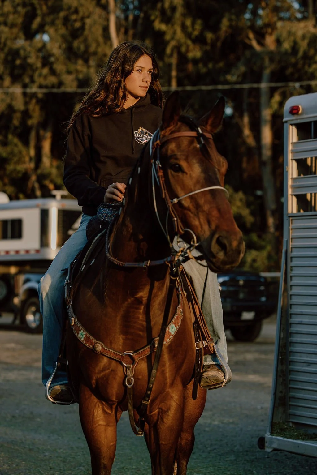 A young woman with long wavy hair riding a brown horse outdoors at sunset.