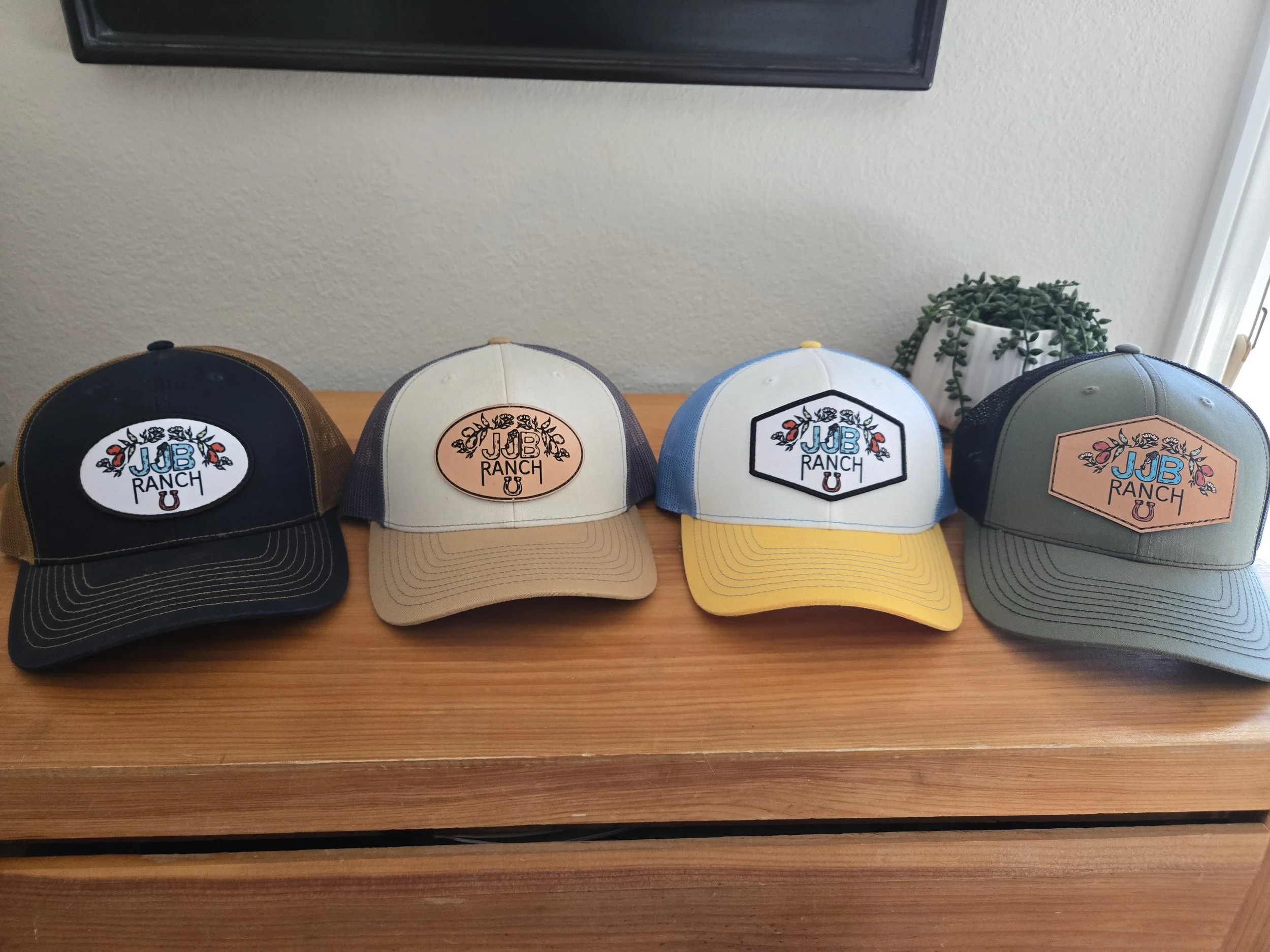 Four baseball caps displayed on a wooden surface. The first cap is black with a colorful embroidered logo reading 'JJB RANCH' with floral and horseshoe motifs. The second cap is beige with a similar logo in a peach-colored patch. The third cap is blue and white with the same logo in a black-bordered hexagon. The fourth cap is gray with the logo in a peach-colored patch.