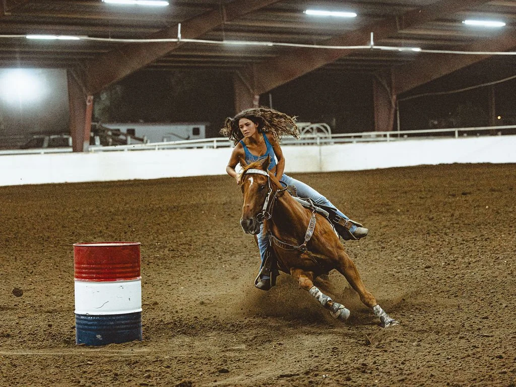 A woman with long curly hair riding a horse around a barrel in an indoor arena for barrel racing.