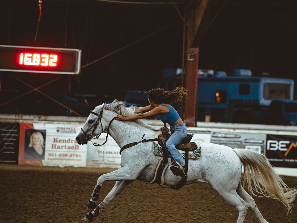 A woman riding a white horse at a rodeo or similar event during the night. The horse and rider are in motion, with the rider leaning forward and gripping the horse's head. There are electronic displays and advertisements in the background.