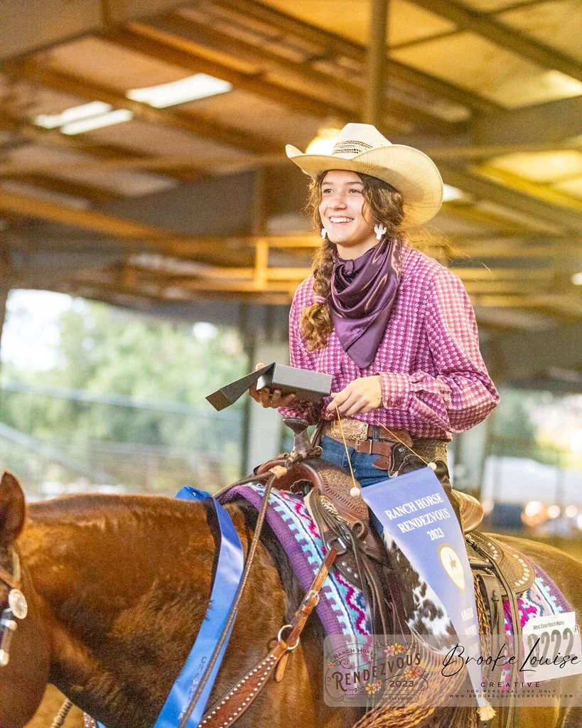 A woman riding a brown horse at a ranch event, wearing a cowboy hat, purple checkered shirt, purple bandana, and jeans, holding a trophy and smiling.