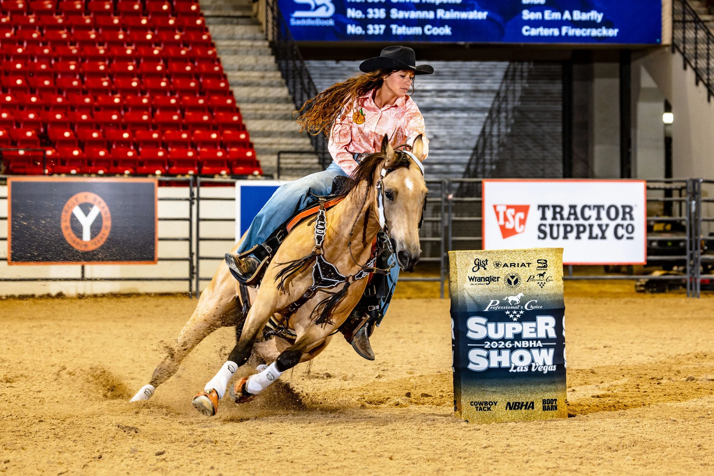 A female rodeo rider wearing a black hat and a western shirt riding a galloping horse during a rodeo event. She is navigating around a barrel marked with event details, inside an arena with empty red stadium seats and sponsor banners in the backgroun