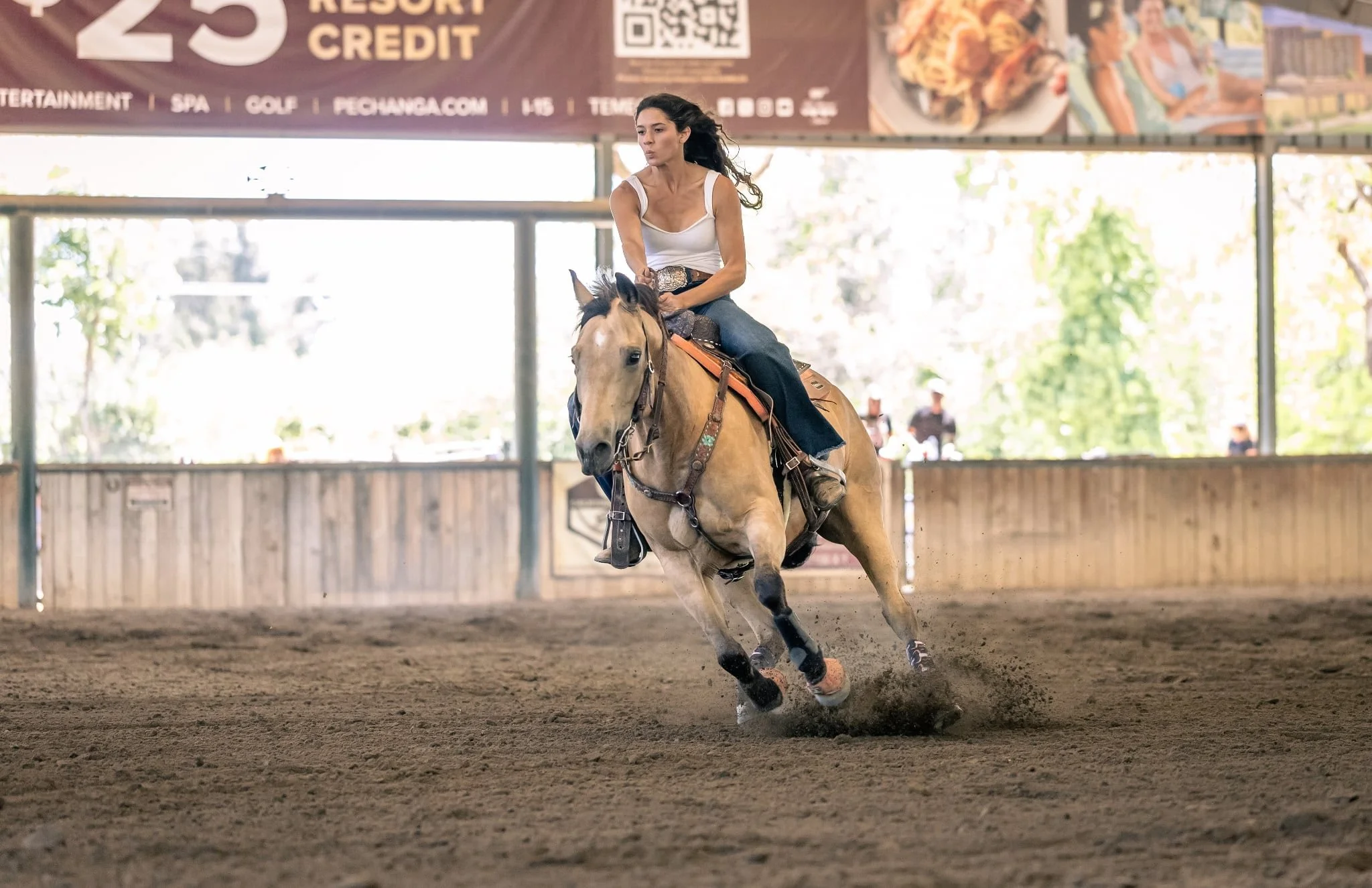 A woman riding a light brown horse with a black mane and tail inside an indoor riding arena. She is wearing a white tank top, blue jeans, and a large belt buckle, with her dark hair flowing as she rides the horse at a gallop. There is a large banner 