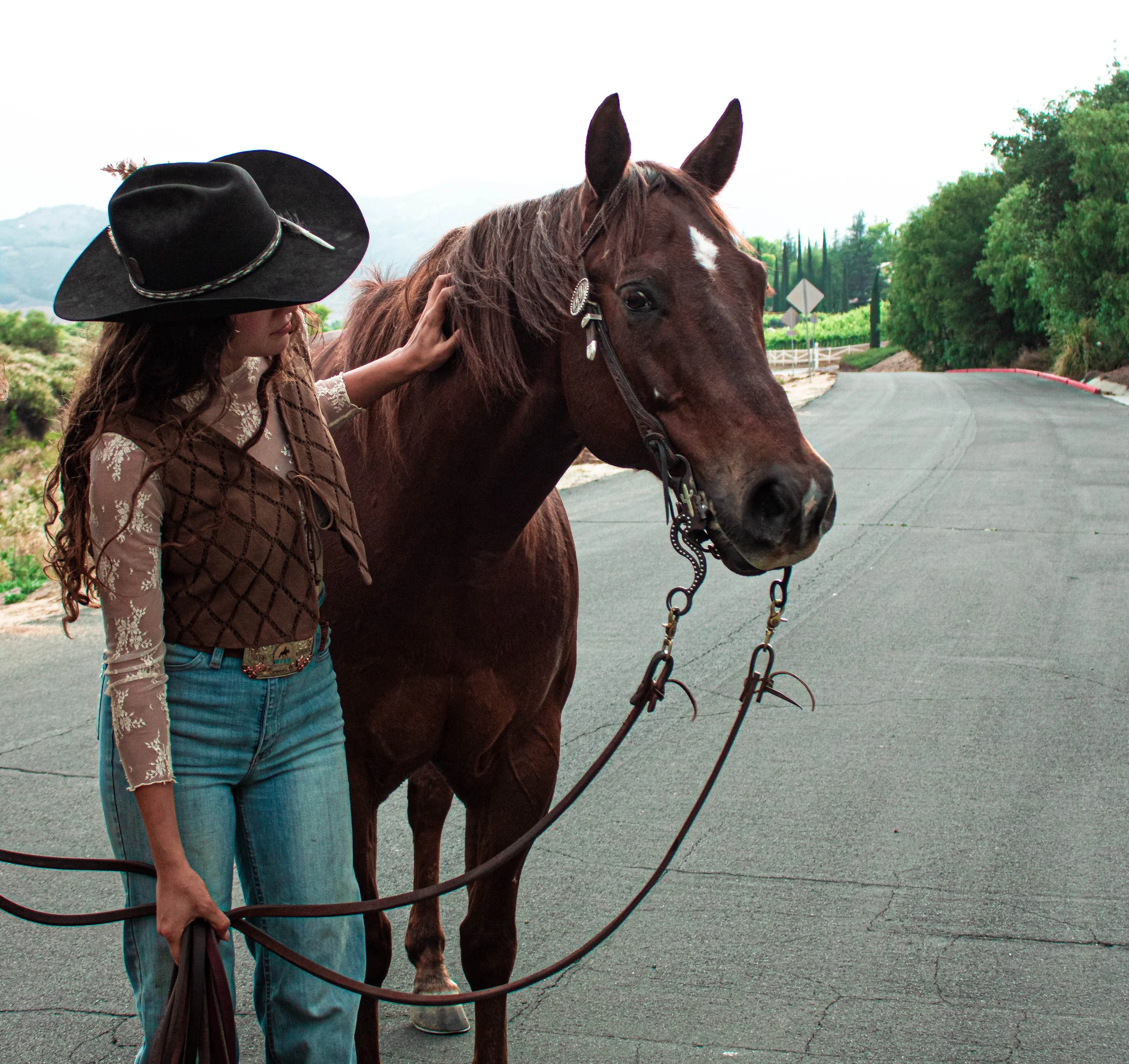 A woman with long curly hair wearing a black cowboy hat, a brown patterned long sleeve shirt, and jeans, petting a brown horse with a white star-shaped marking on its forehead on a paved road with greenery in the background.