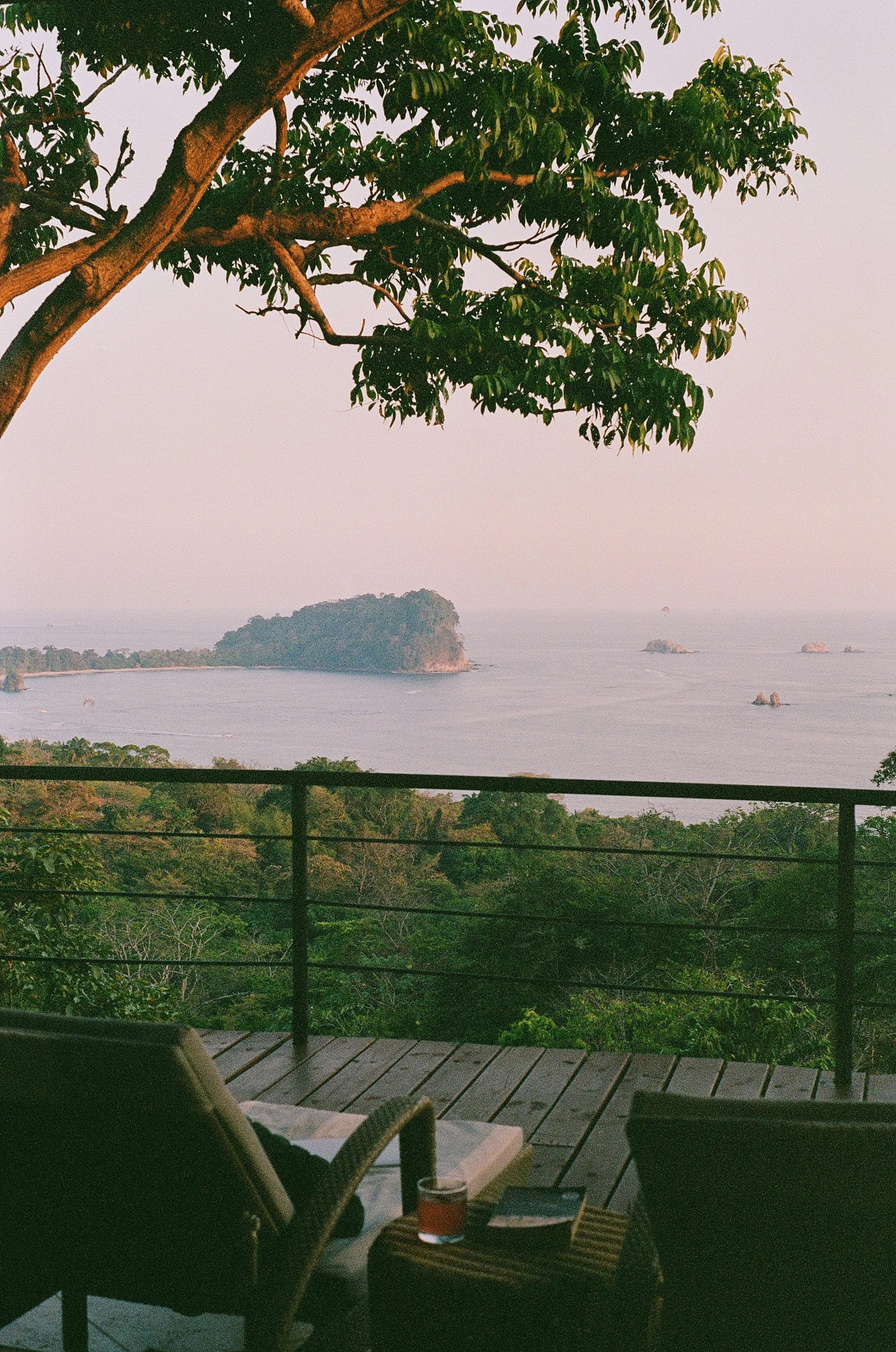 A cozy outdoor patio area overlooking the ocean with lush greenery in the foreground. There is a wooden deck with chairs, a table with a drink and some books, and a view of a small island and rocks in the distance at sunset or dusk.