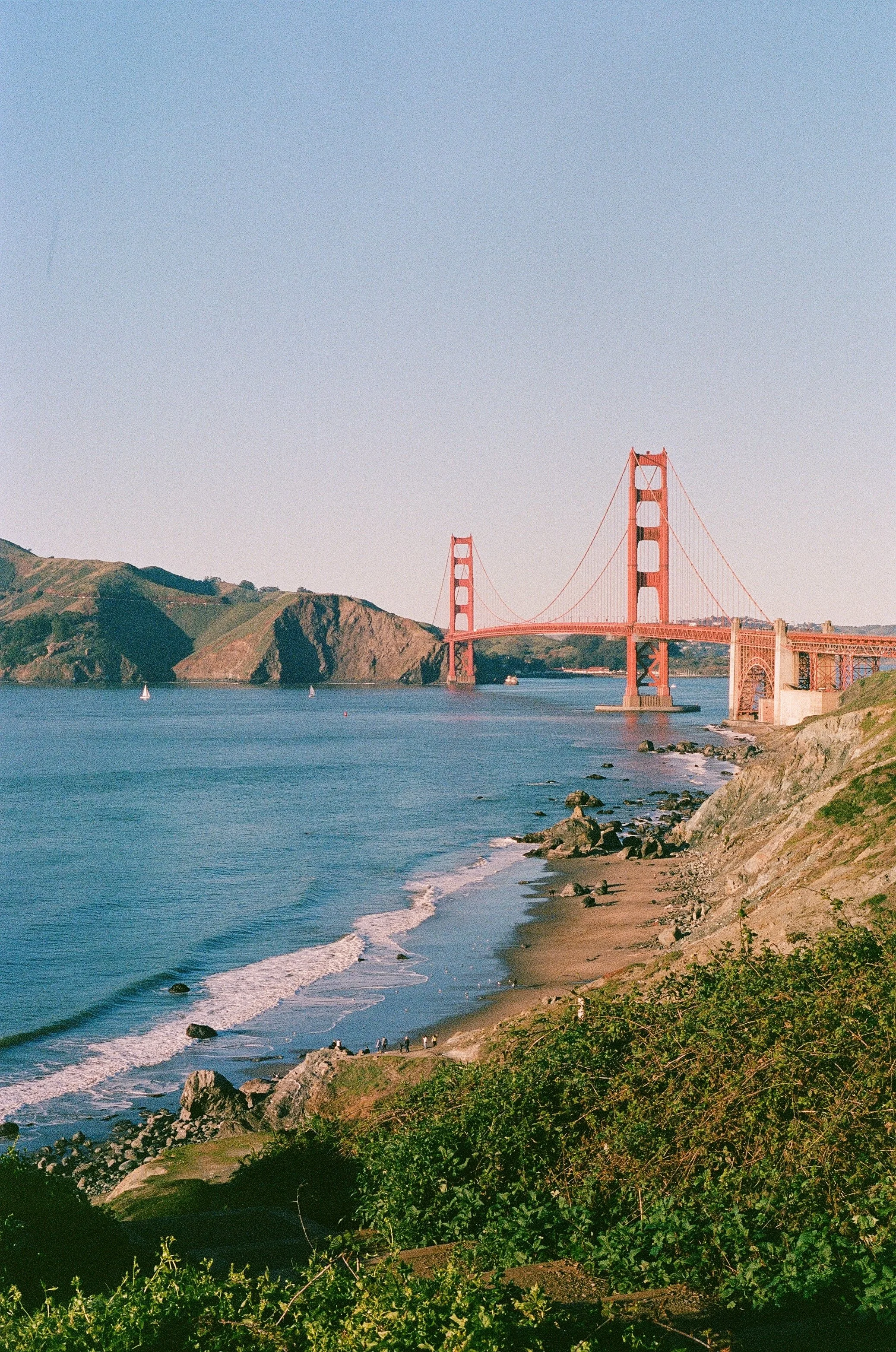 The Golden Gate Bridge in San Francisco, California, spans across the water with hills in the background during daylight. There are sailboats on the water, and people walking along the rocky shoreline.