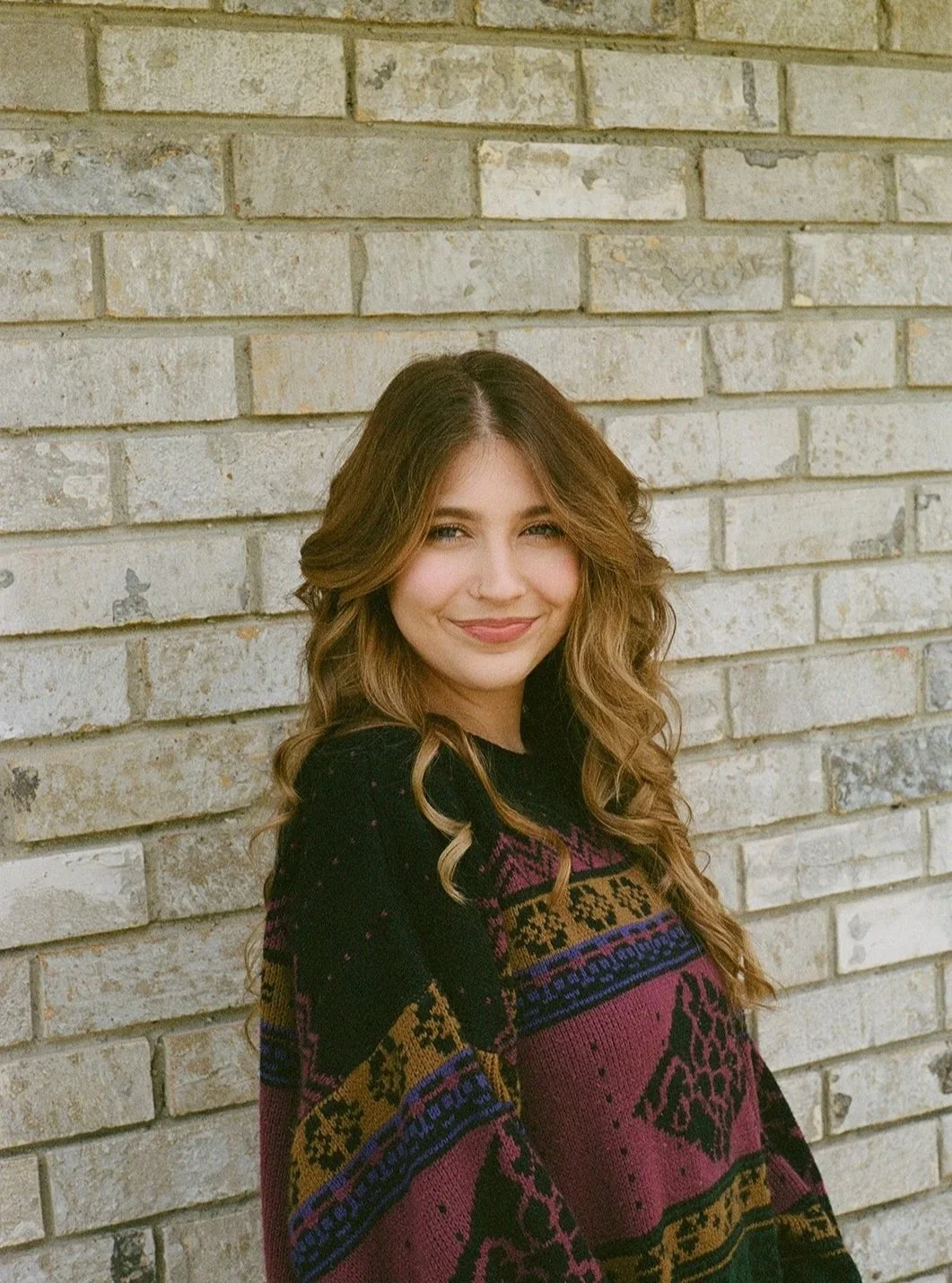 A young woman with long, wavy brown hair smiling and standing against a beige brick wall. She is wearing a colorful, patterned sweater with animal and geometric designs.