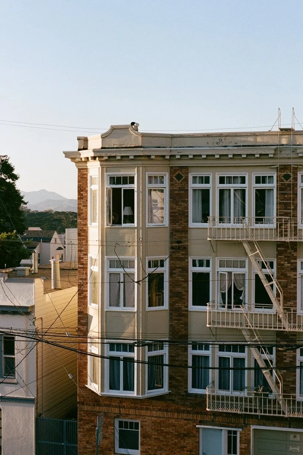 A multi-story residential building with bay windows and fire escapes, with the sky and some distant mountains in the background.