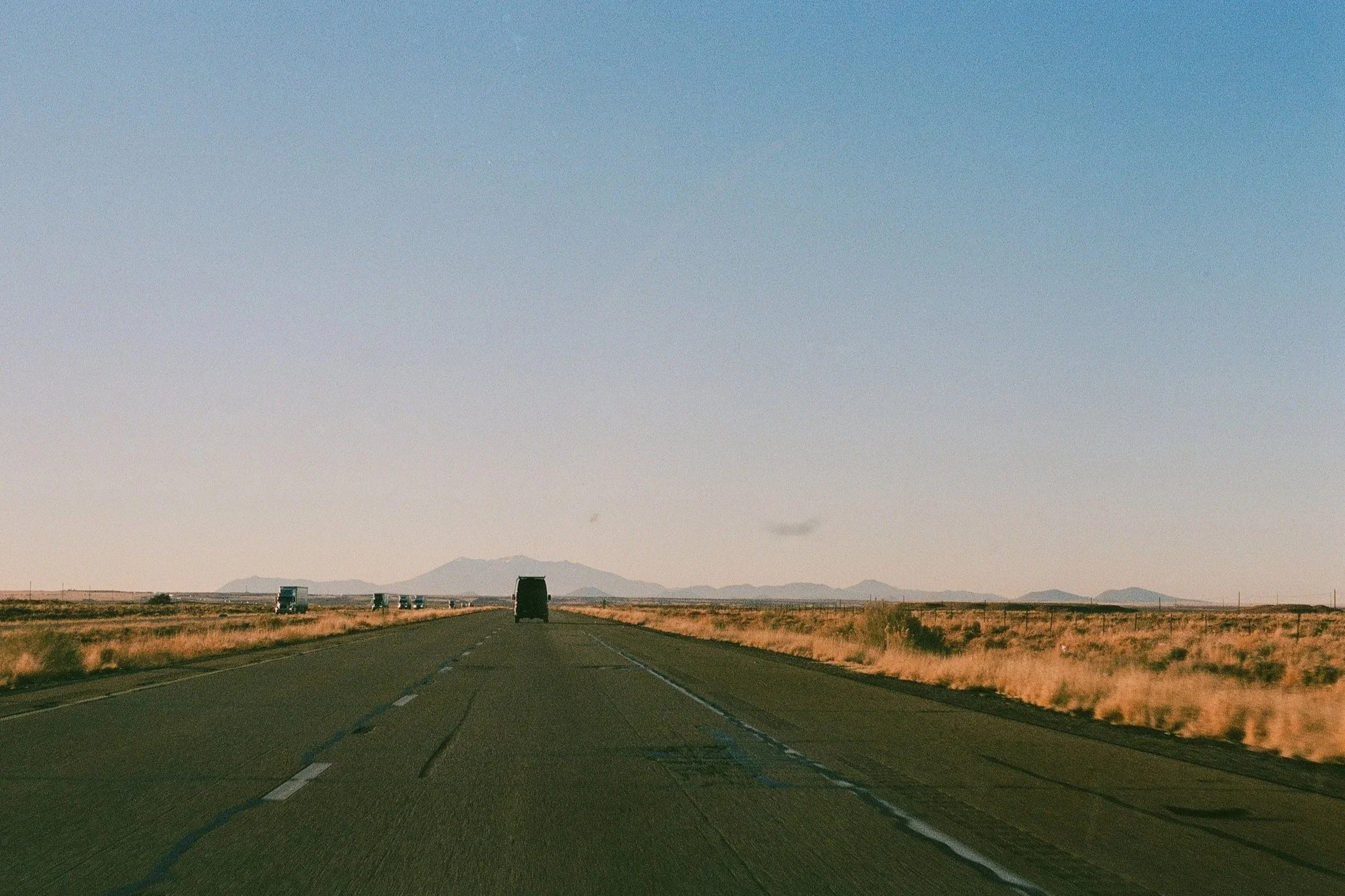 A straight highway in a flat rural area with multiple vehicles traveling in both directions, distant mountains in the background, and a clear sky overhead.