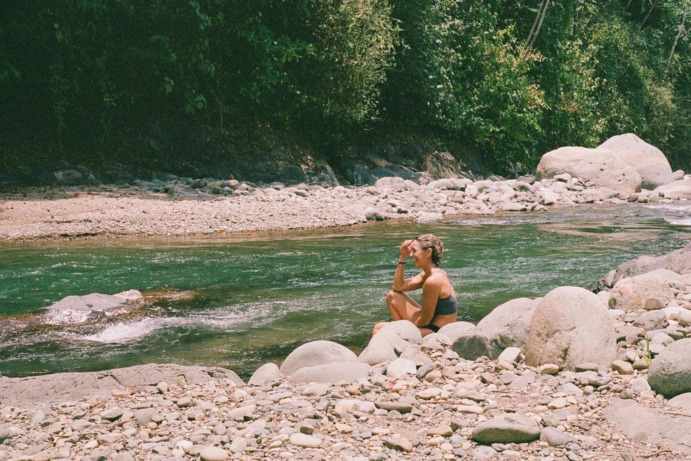 A woman sitting on large rocks by a river, with forested area and rocks in the background.