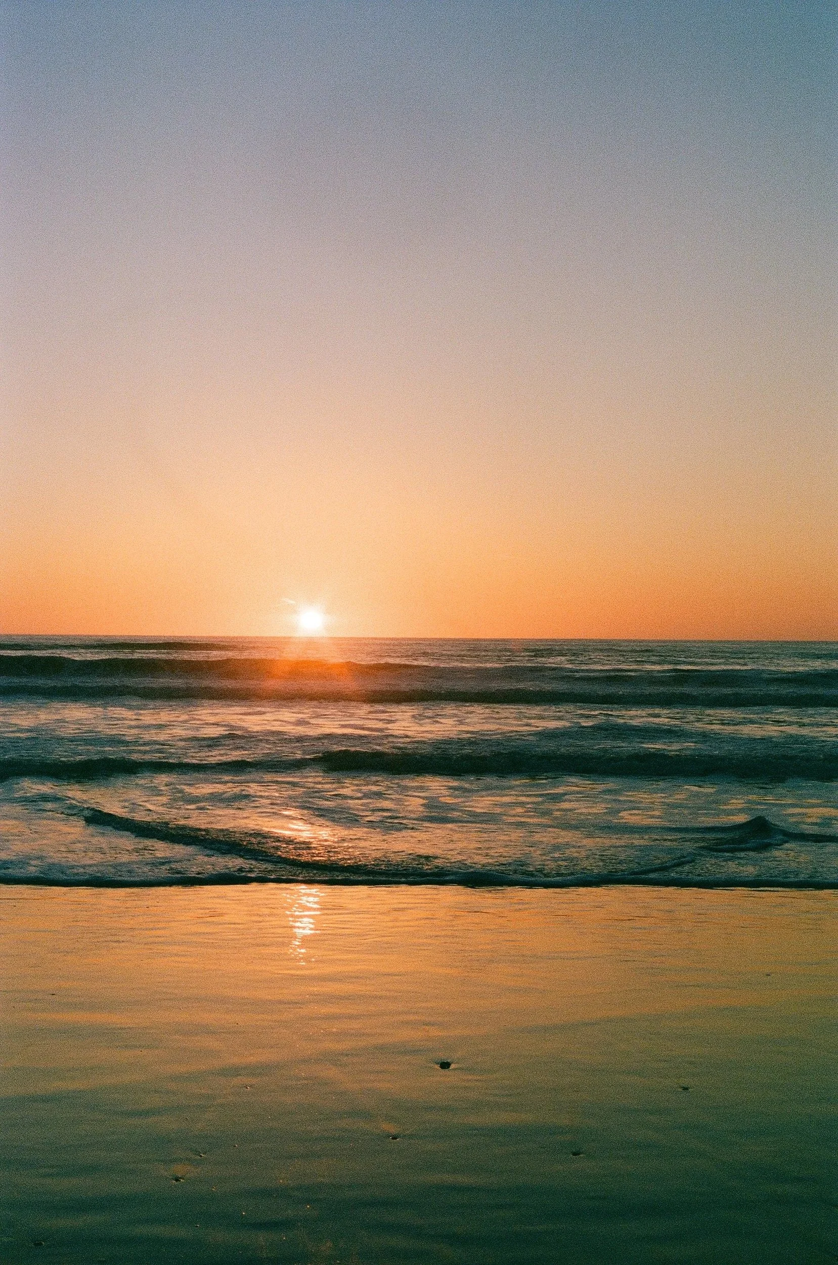 Sunset over the ocean with the sun near the horizon, casting reflections on the water and gentle waves along the shore.