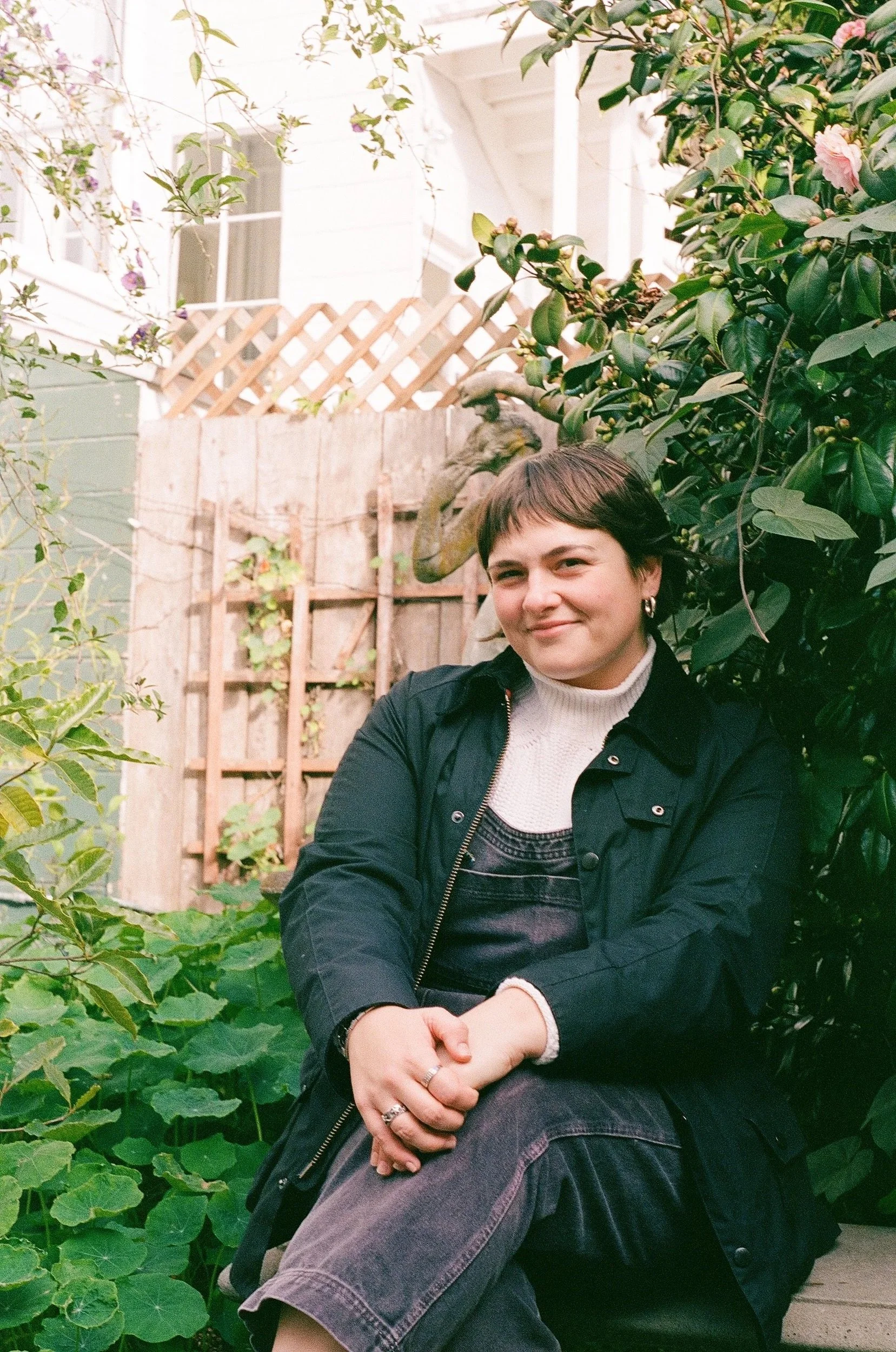 A woman with short brown hair wearing a black jacket and white turtleneck, seated outdoors among green plants and bushes, with a wooden fence and a house in the background, smiling at the camera.