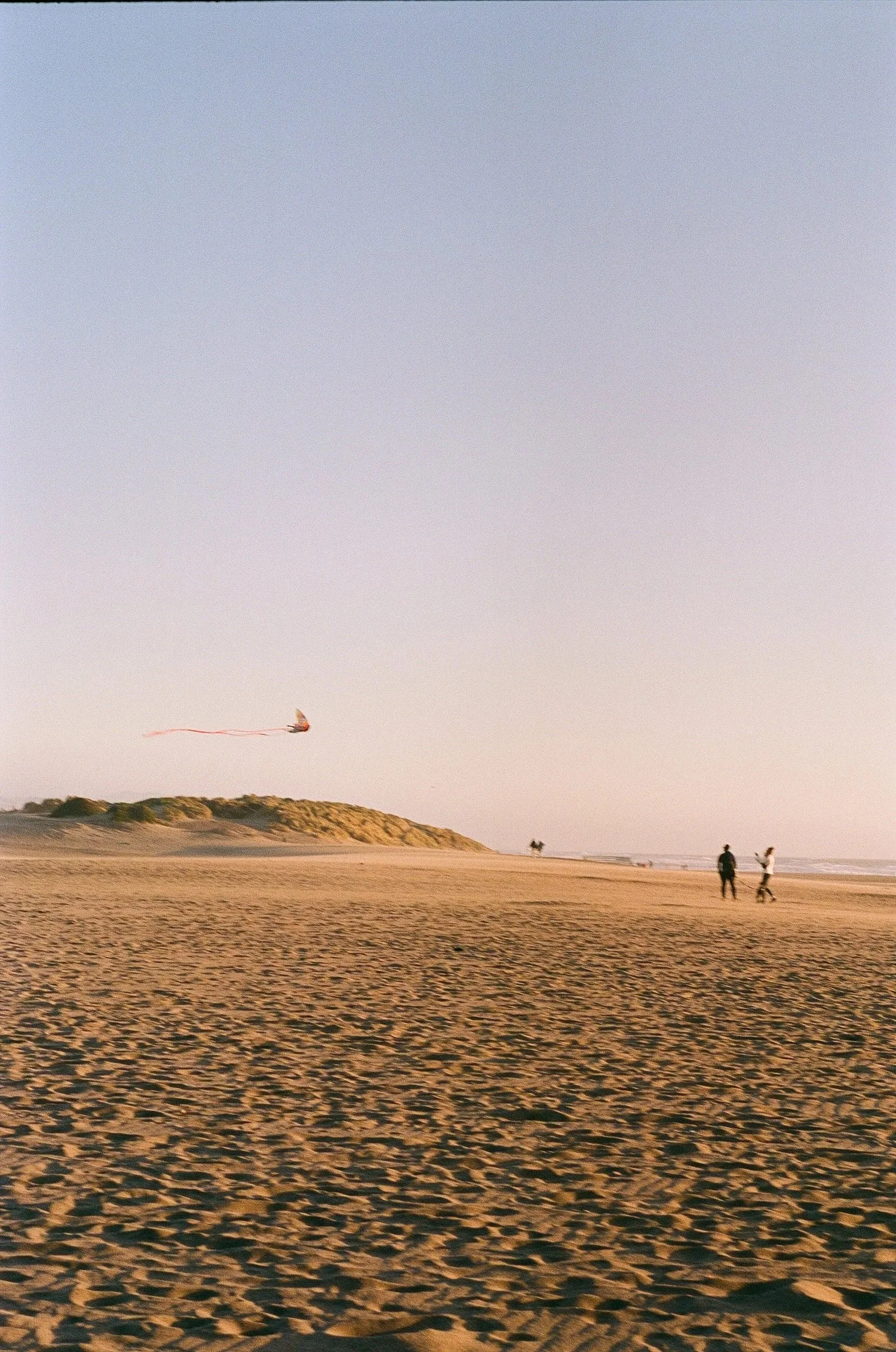 A sandy beach at sunset with a kite in the sky and four people walking along the shoreline.