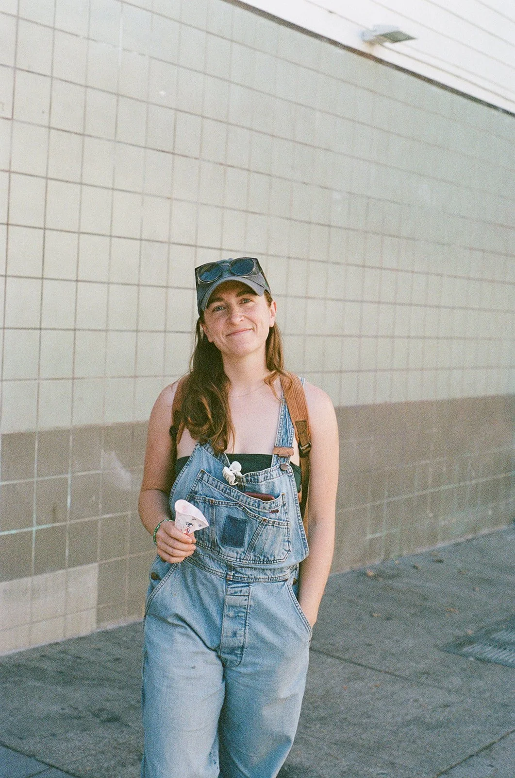 A young woman with long brown hair, wearing a black tank top, denim overalls, a gray baseball cap with sunglasses on top, and a brown backpack. She is holding a small ice cream cone and smiling, standing against a tiled wall outdoors.