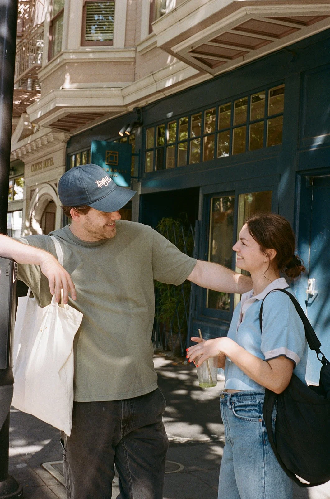 A young man and woman are smiling and talking on a city street, with the man leaning on a parking meter and the woman holding a drink. They appear happy and engaged in conversation.