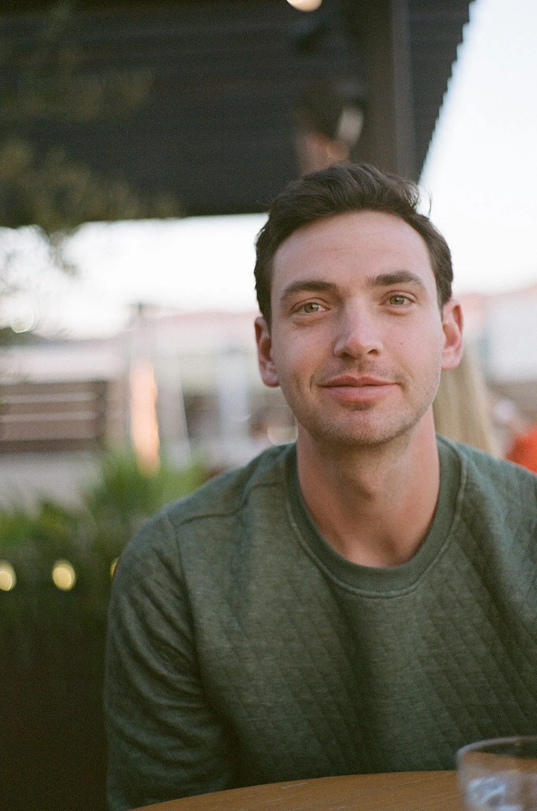 A young man with dark hair and light skin, wearing a dark green sweatshirt, sitting outdoors at a table with a glass of water, smiling softly.