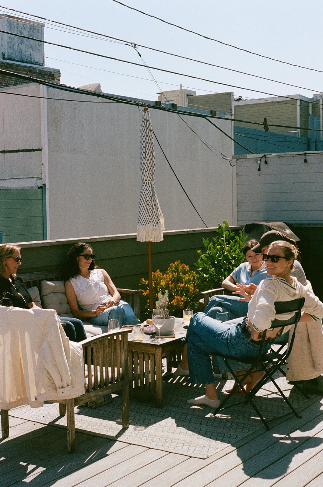 Group of five women sitting and chatting on a patio, with outdoor furniture, drinks, and a closed umbrella under a sunny sky.